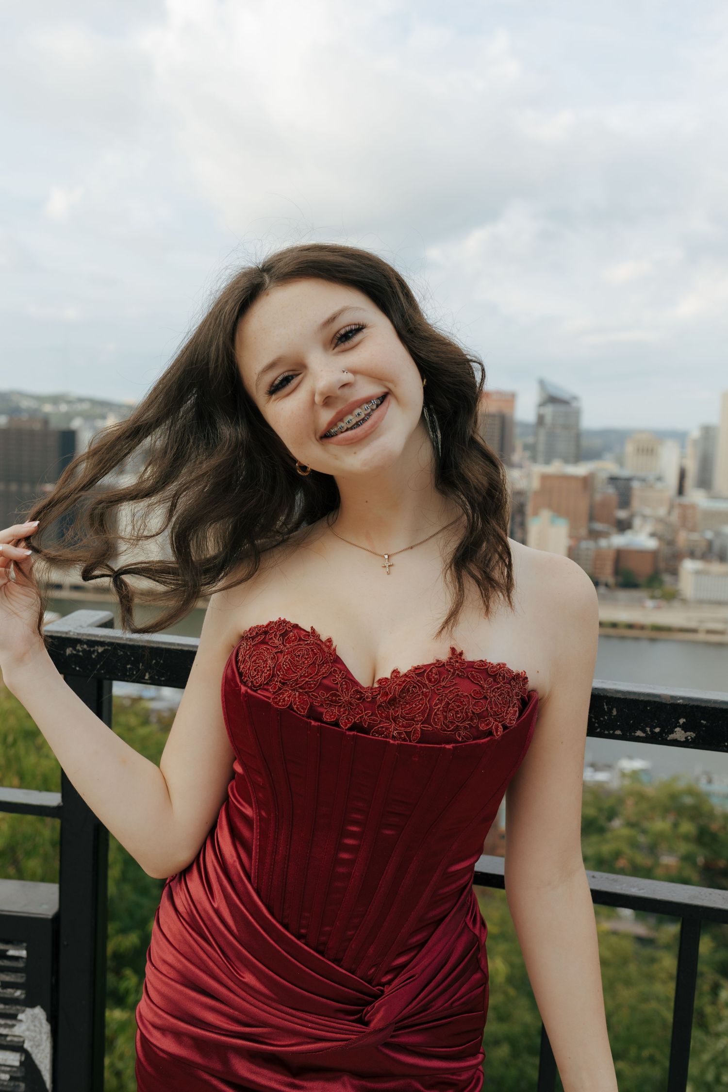 Young girl in red strapless gown smiles with city skyline backdrop at outdoor overlook for her homecoming at Downtown Pittsburgh overlook