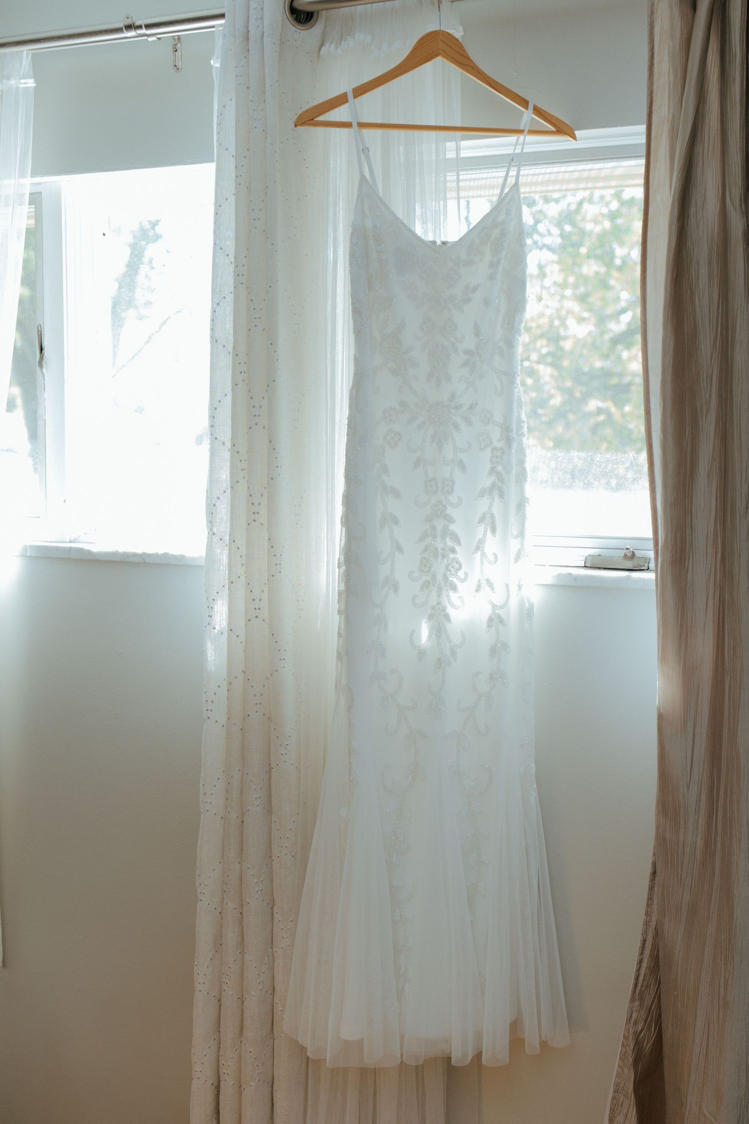 Delicate white lace wedding dress hanging on a wooden hanger in a sunlit window before the ceremony.
