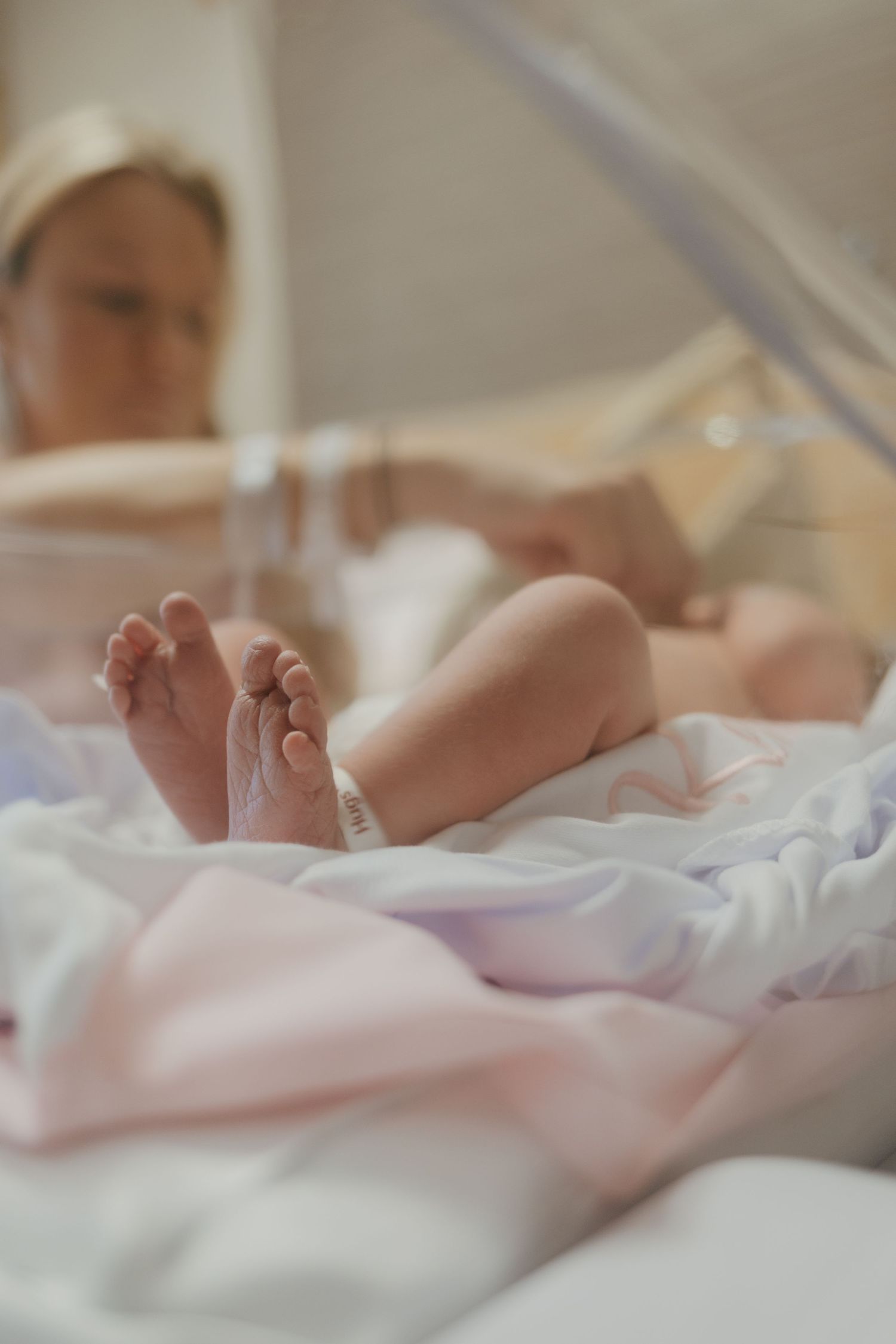 Newborn baby feet with hospital ID bracelet in a clear bassinet, with mother resting in the background.