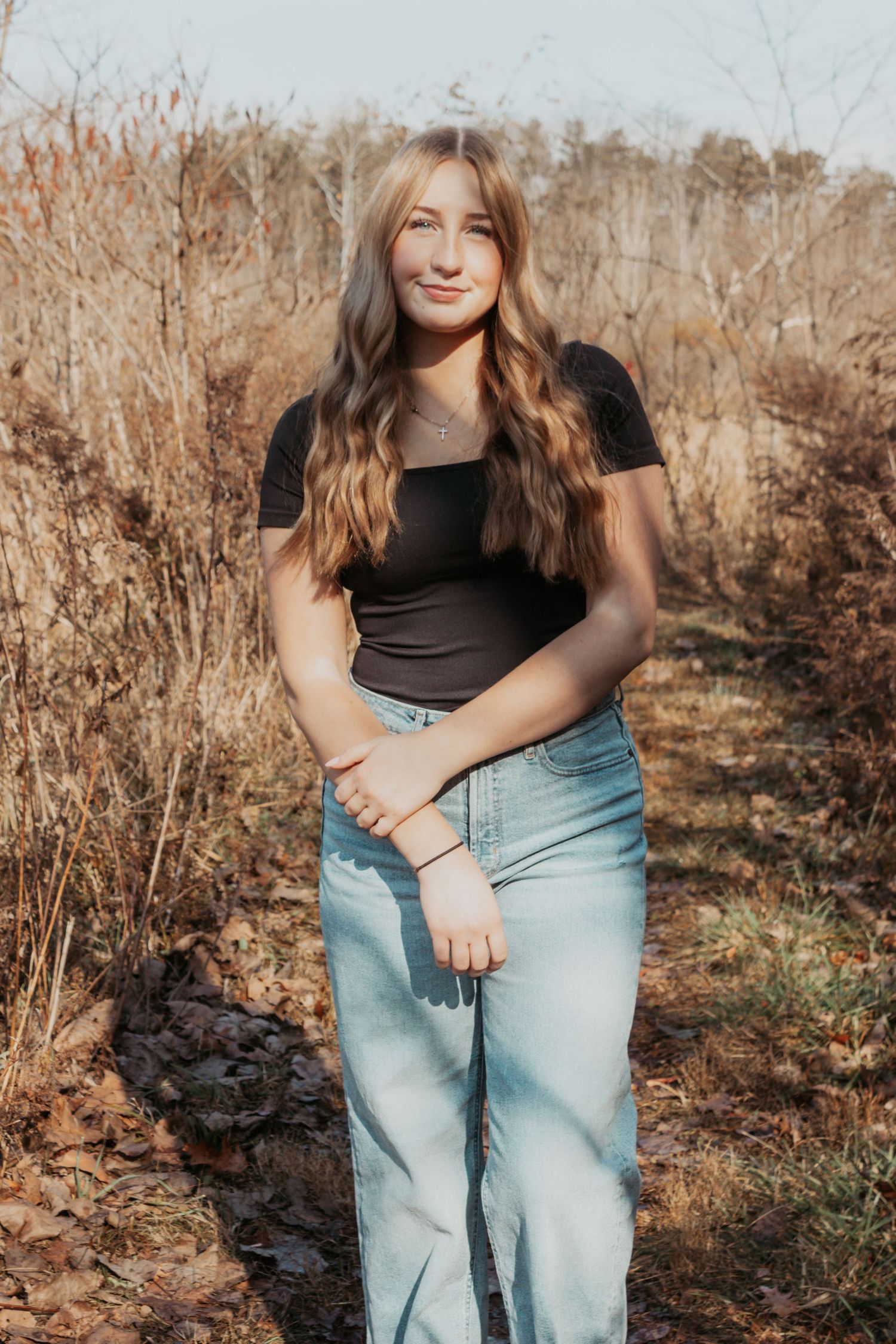Young woman with long wavy hair in black top and jeans posing in autumn field.