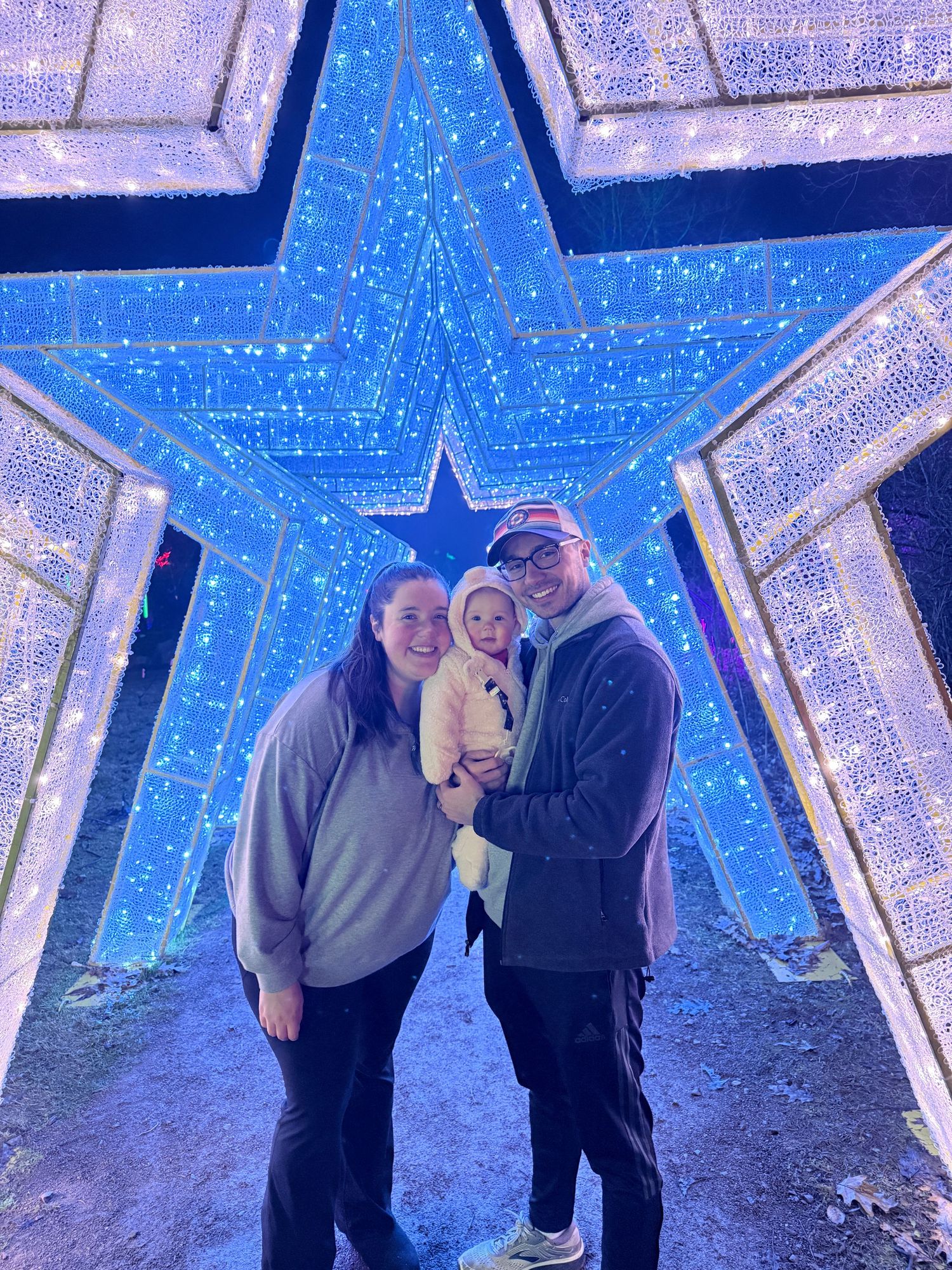 Family of three smiling under blue star light display at holiday lights event.