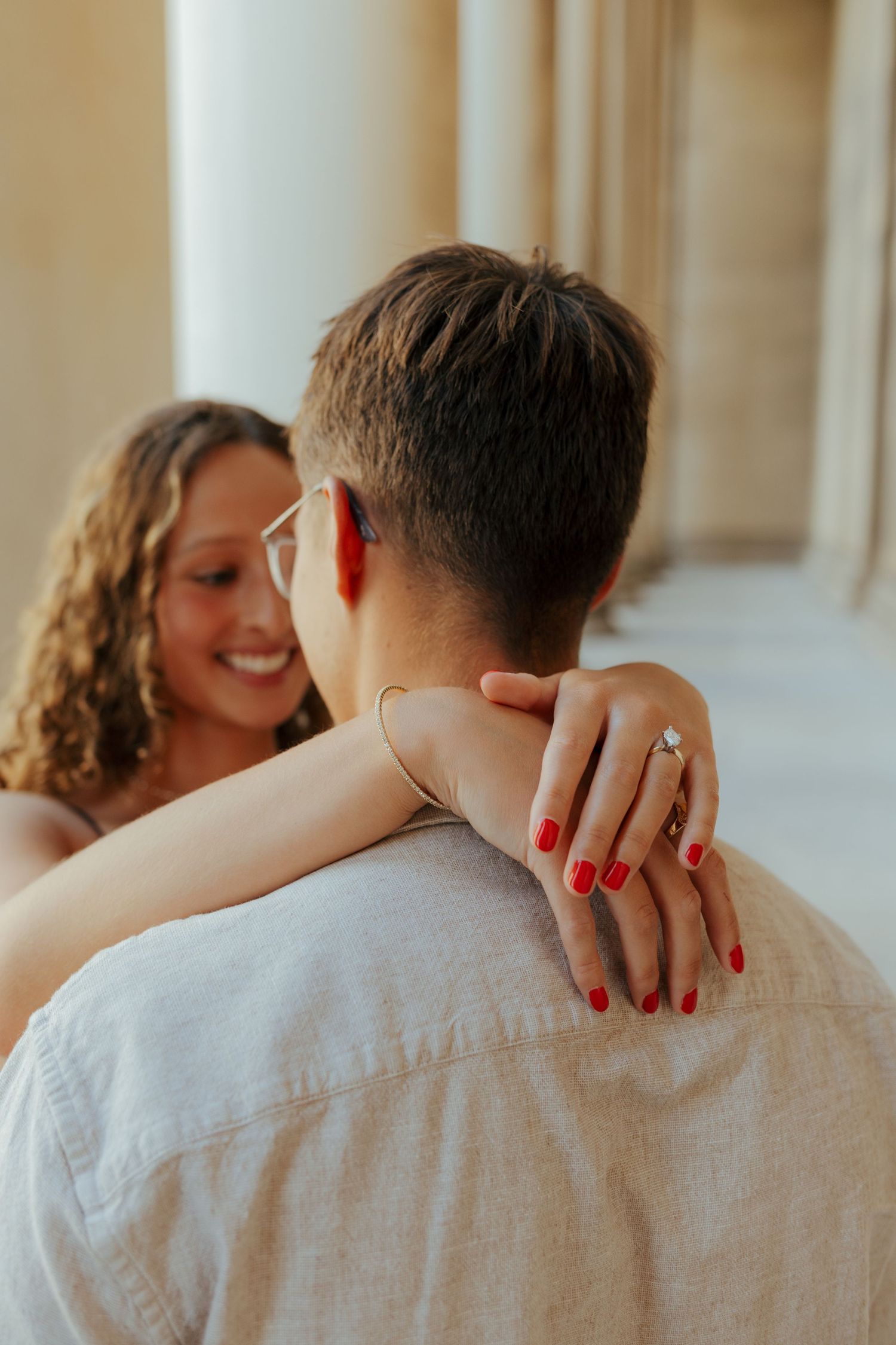 Smiling woman with curly hair embracing man from behind showing engagement ring with red nails.