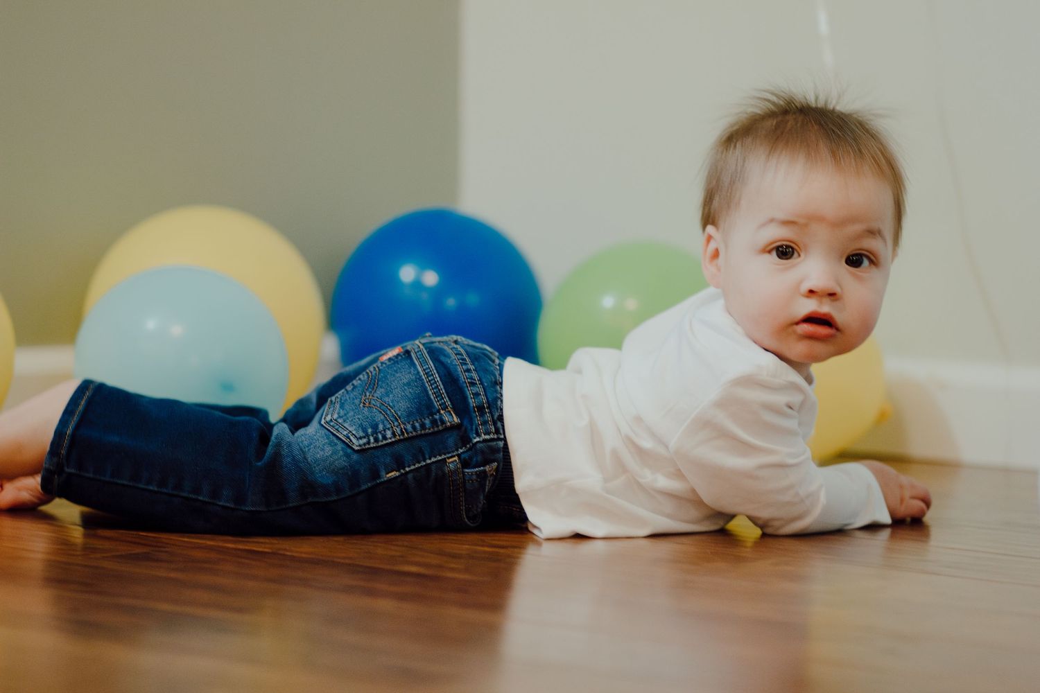 Baby with Down syndrome crawling on hardwood floor surrounded by colorful balloons at birthday party.