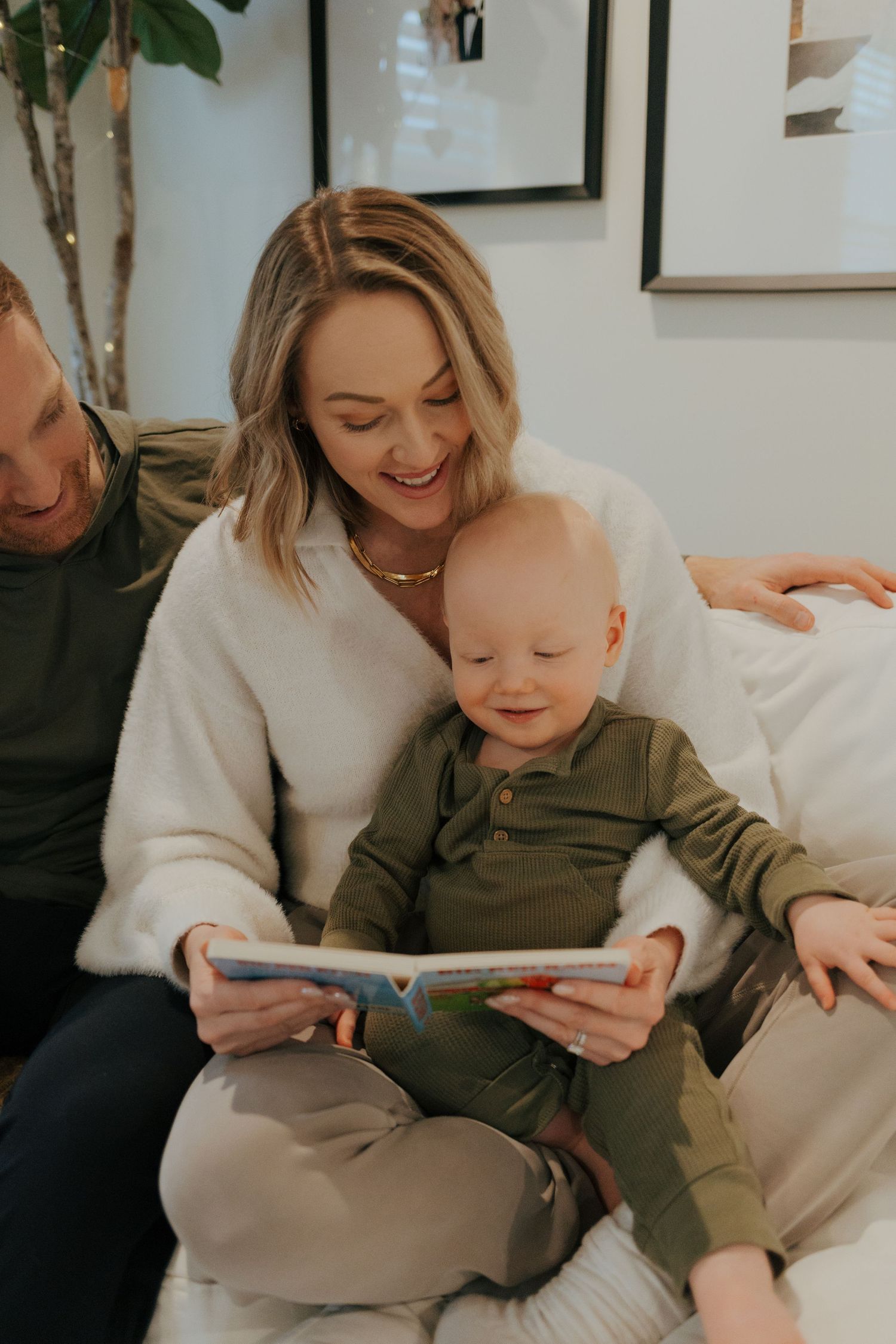 Parents laugh with happy baby while reading a colorful picture book together on a cozy couch.