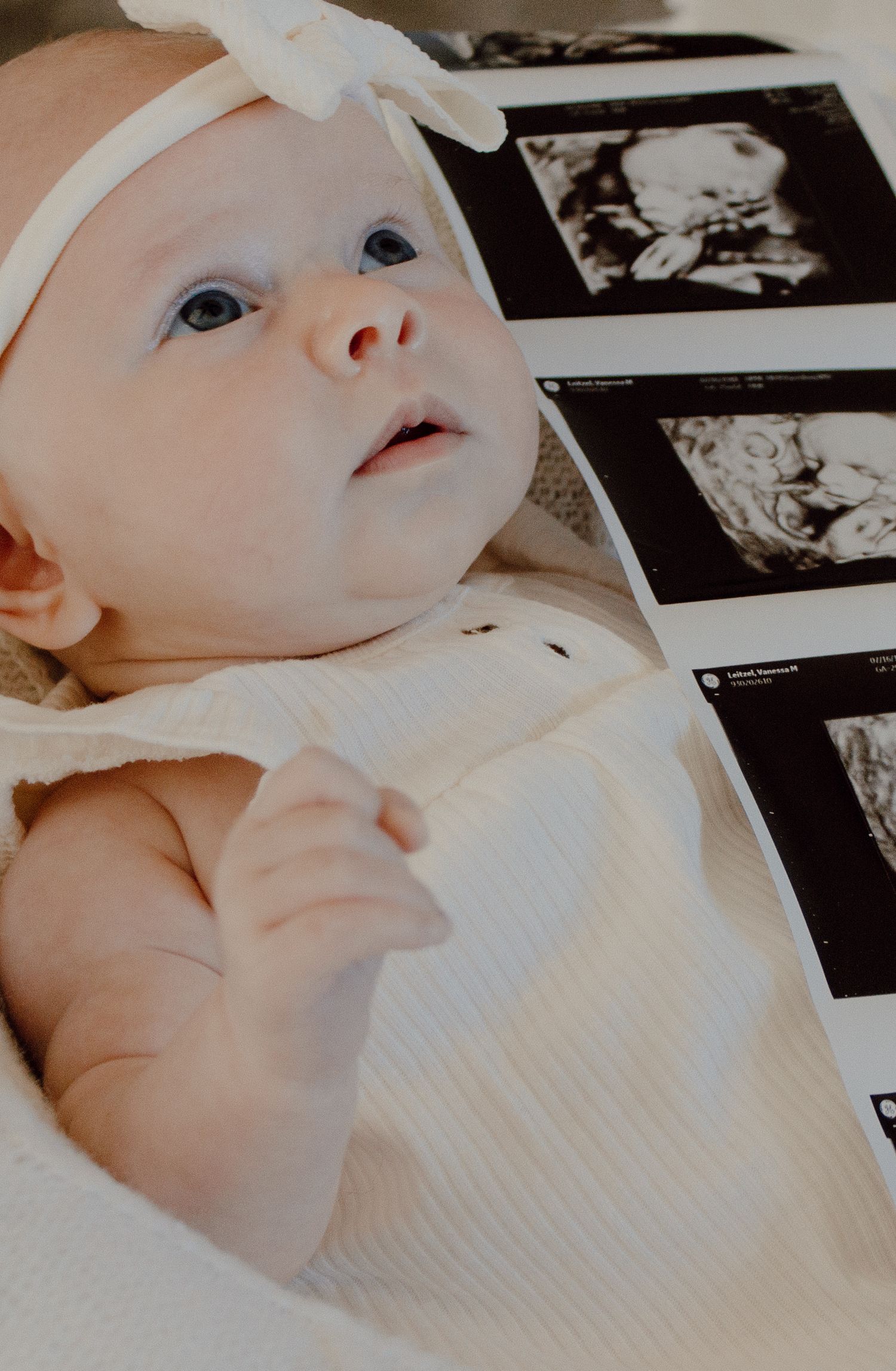 Adorable newborn baby girl in white bow headband lying beside ultrasound sonogram photos.