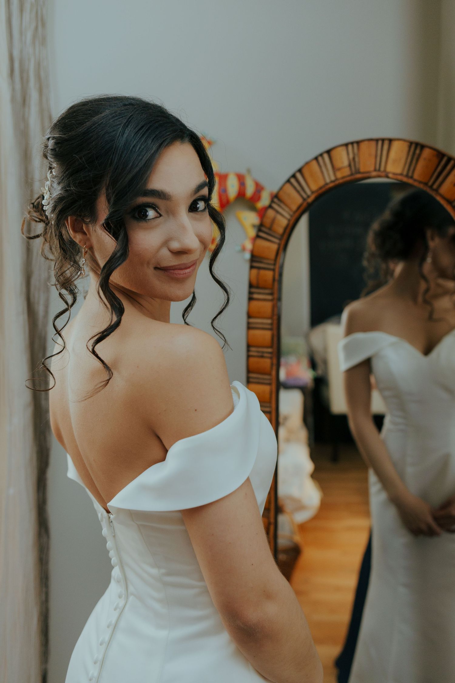 Beautiful bride in off-shoulder white wedding gown smiling over her shoulder near a mirror.