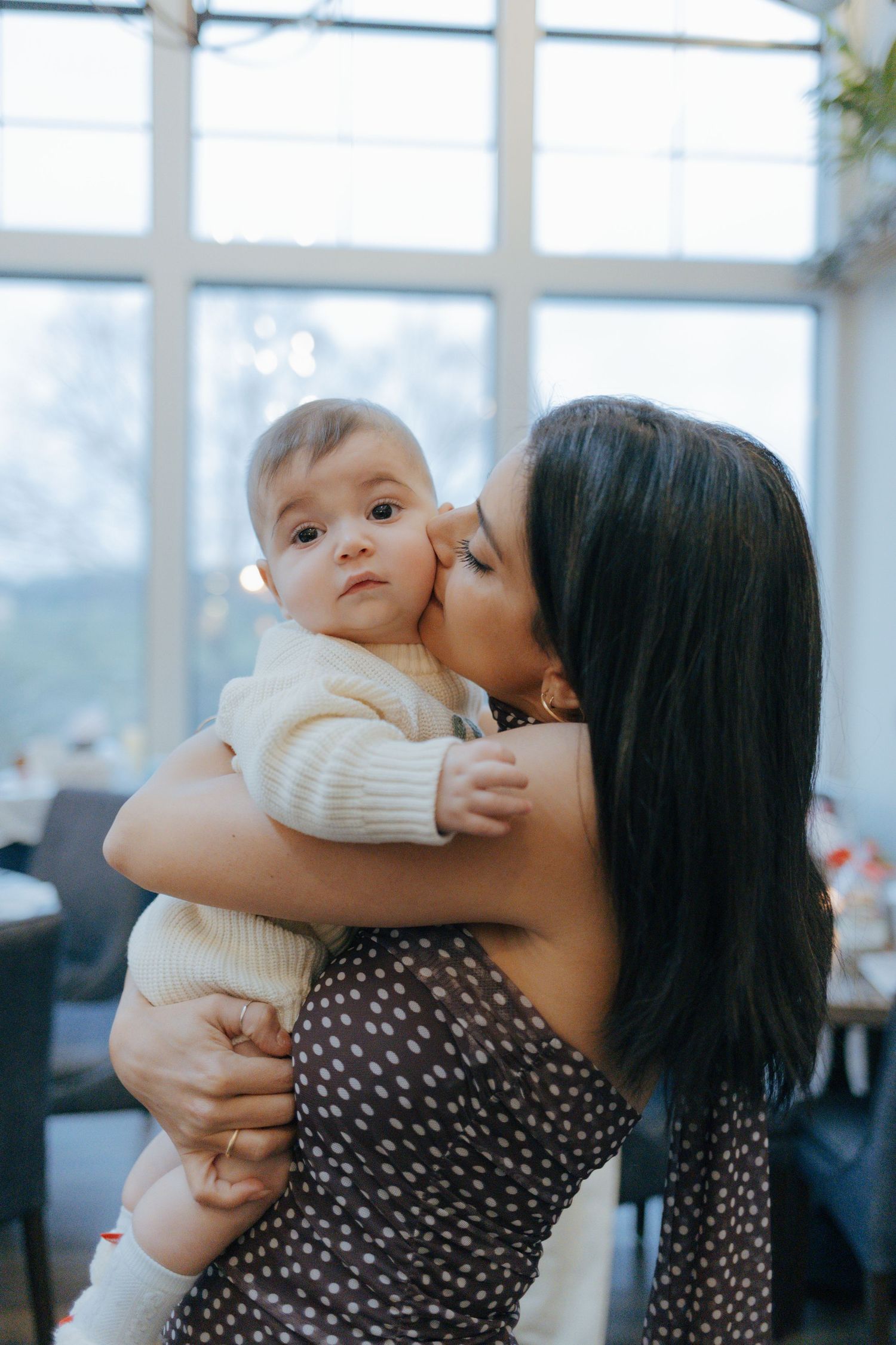 A dark-haired woman in a floral dress tenderly kisses a baby on the cheek in a bright, windowed room.