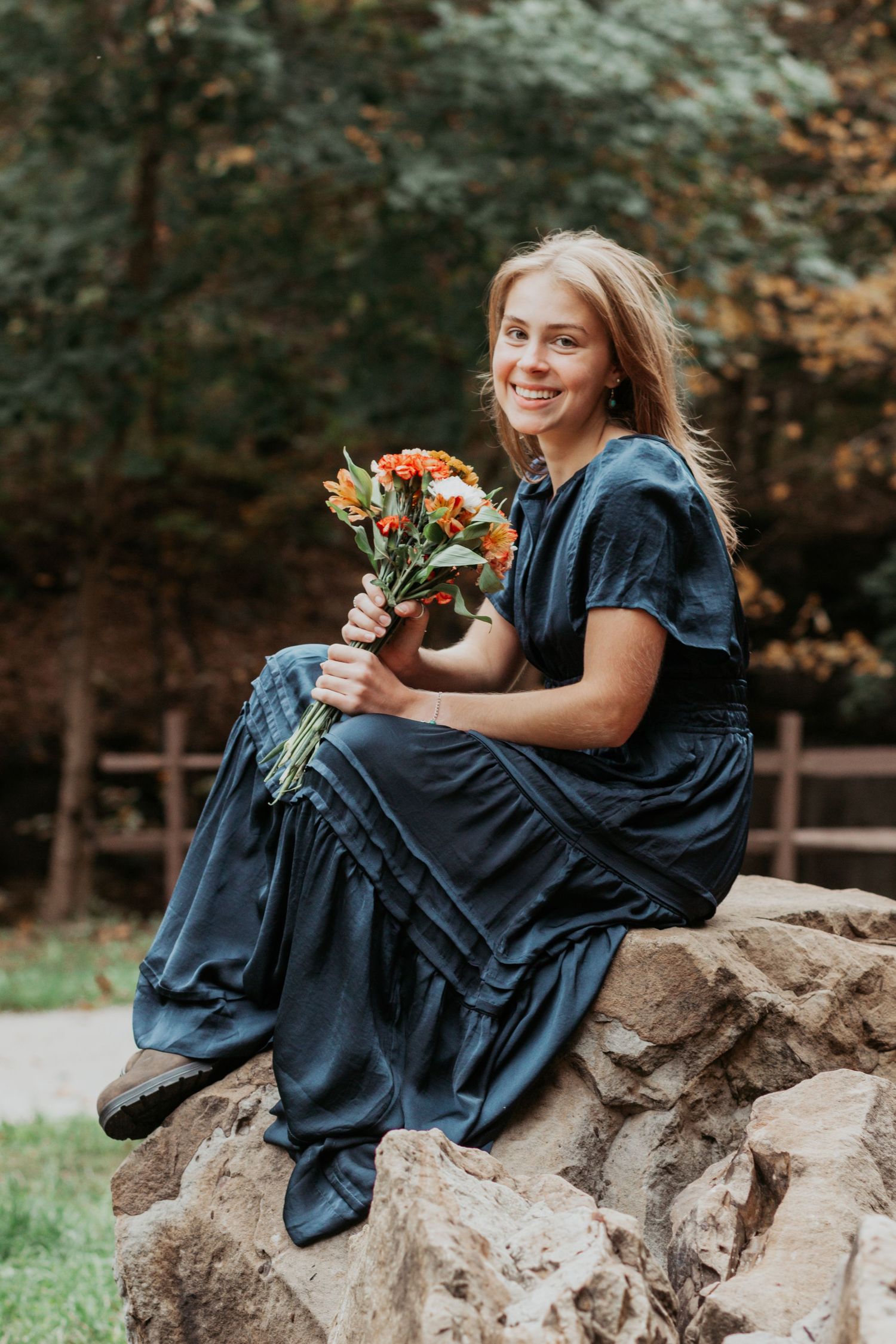 Smiling blonde woman in navy dress holding orange flower bouquet on rocks outdoors.