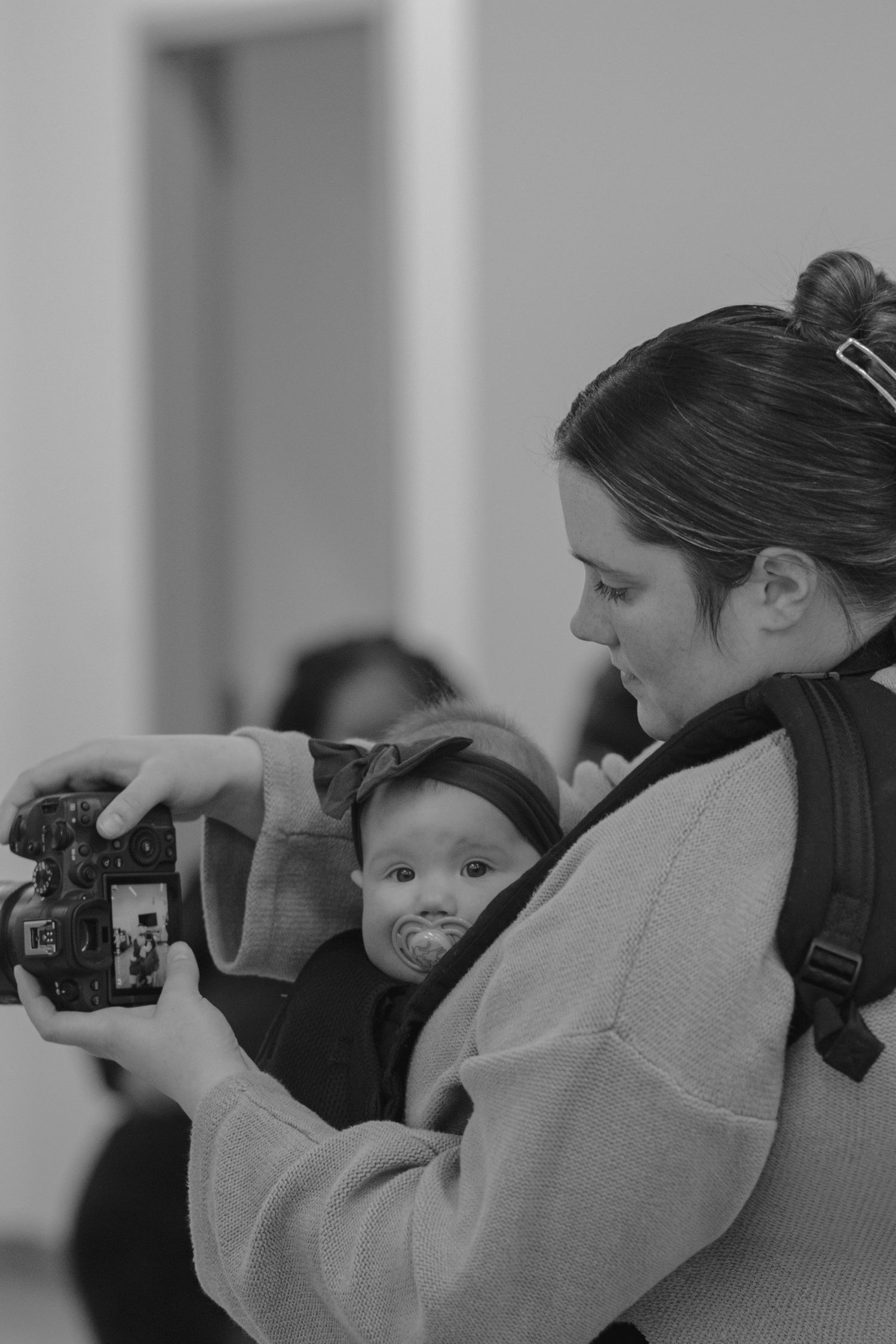 Black and white photo of a mother carrying baby in carrier while taking photos with DSLR camera. Laced In Purpose Photography.