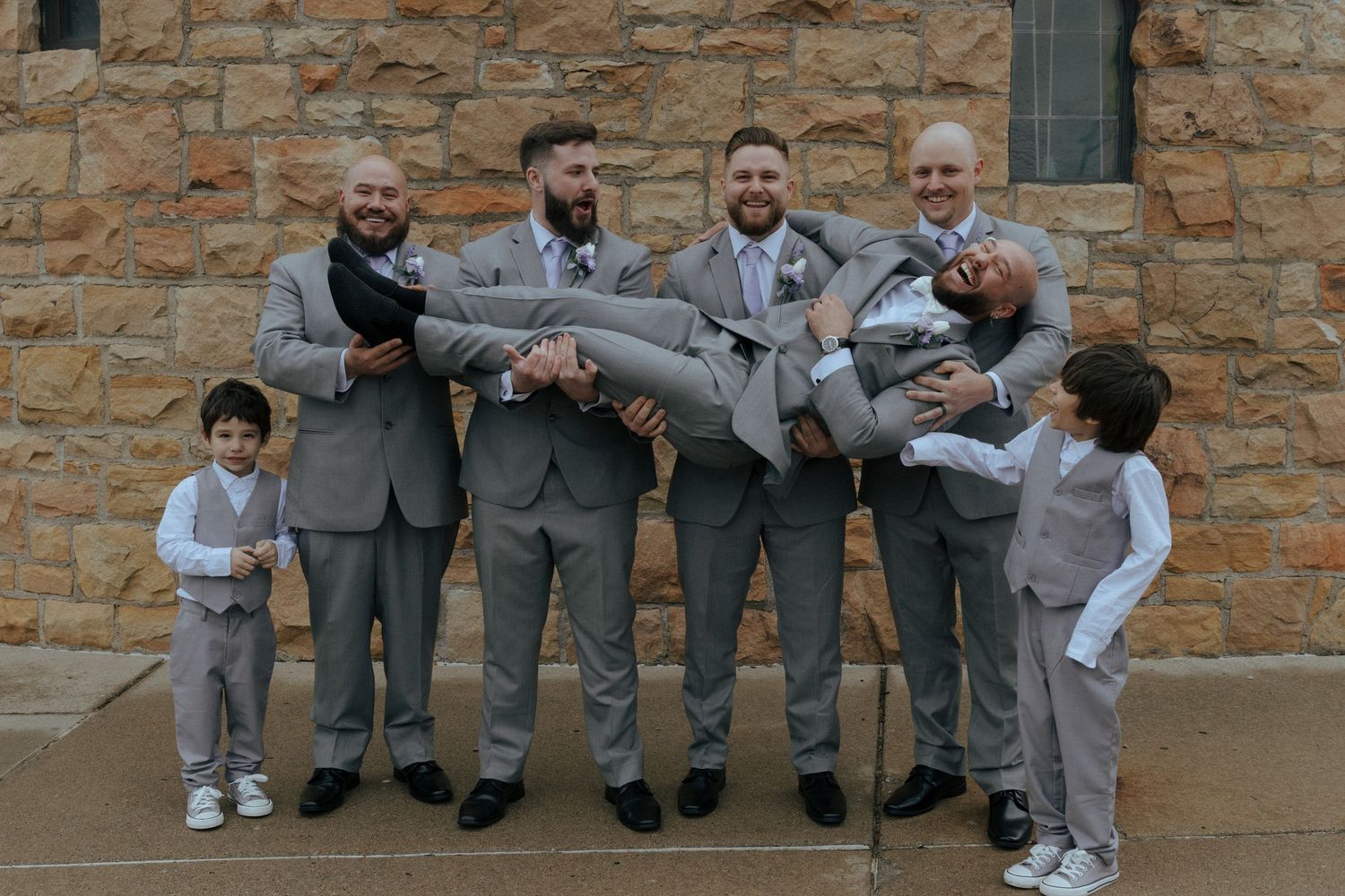 Groomsmen in gray suits playfully carrying the groom outside a rustic stone building with two ring bearers.