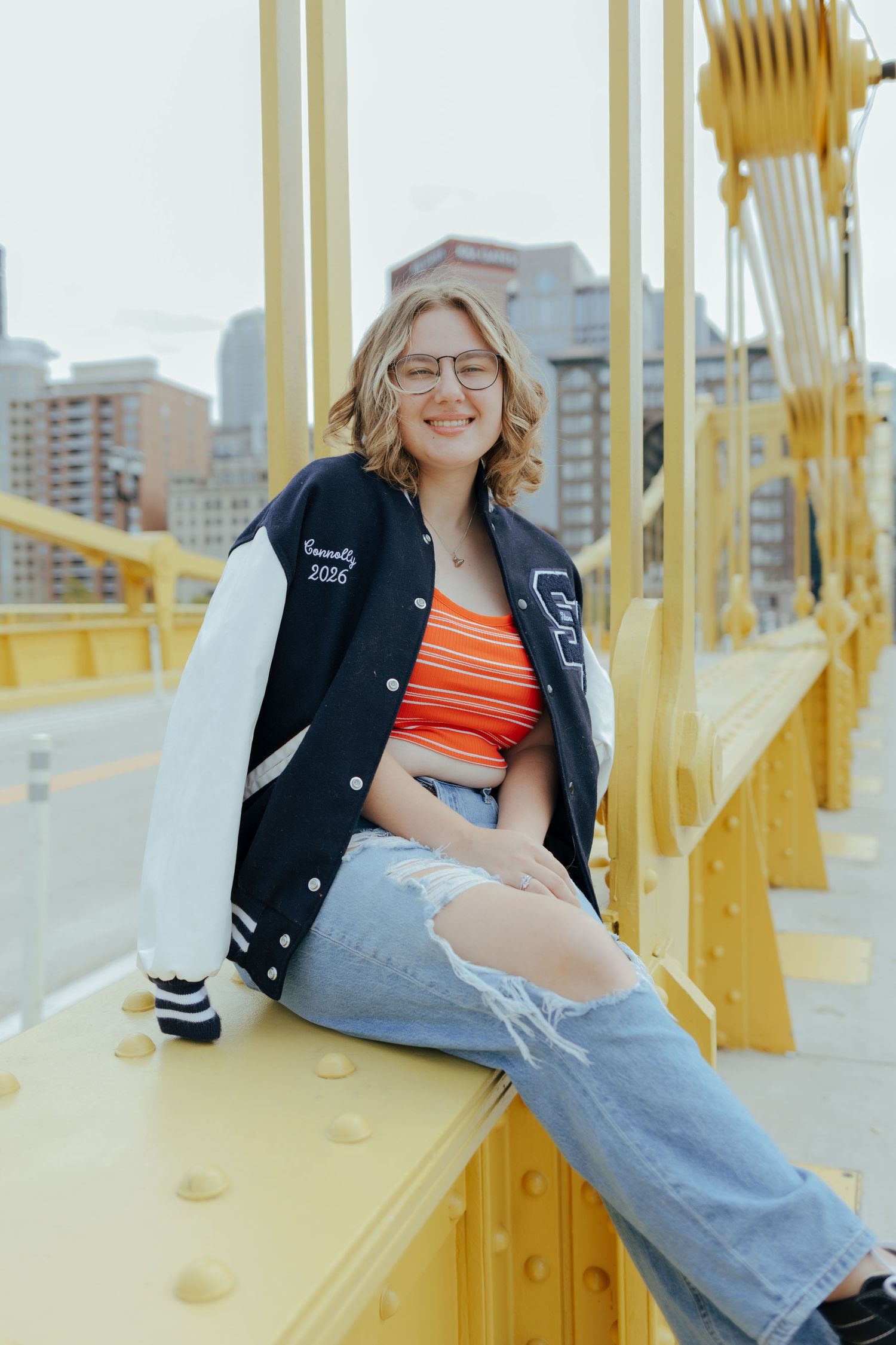 Smiling teen in varsity jacket and ripped jeans sits on yellow bridge with city skyline backdrop.