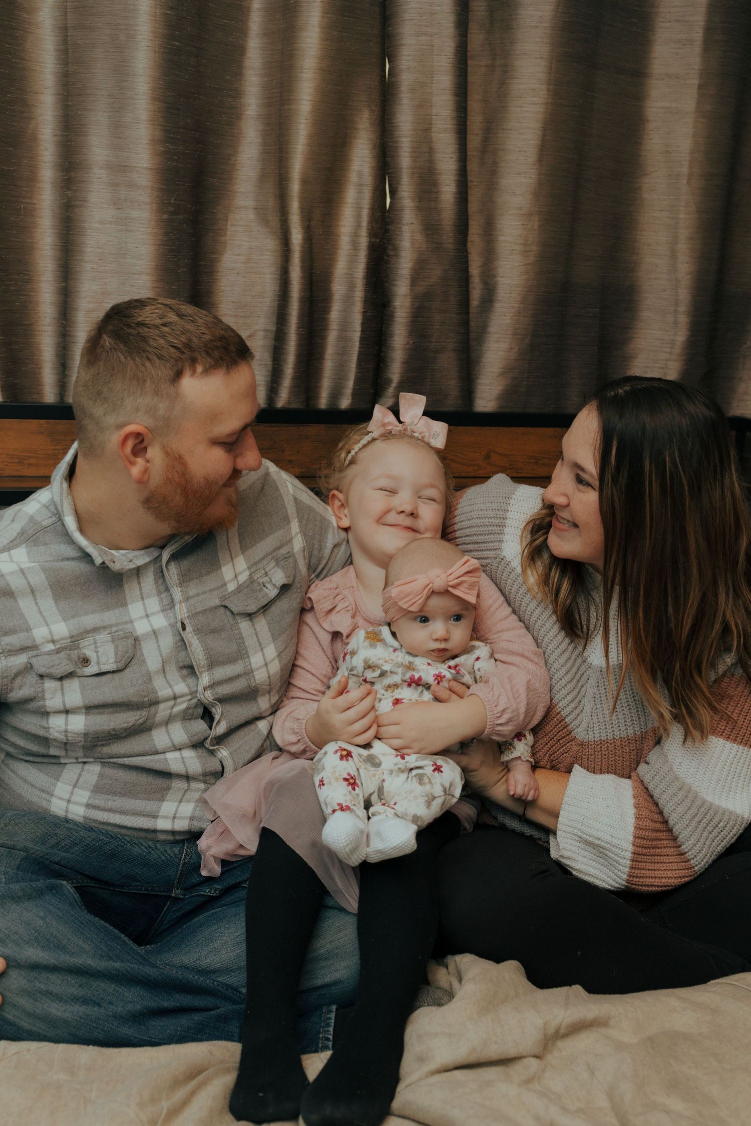 Smiling family of four with parents and two young daughters sitting together on a bed.