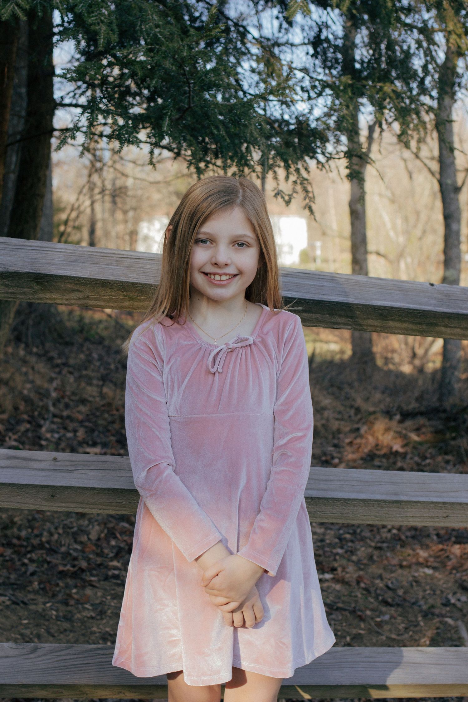 Smiling girl in pink velvet long-sleeve dress stands in front of a rustic wooden fence outdoors.