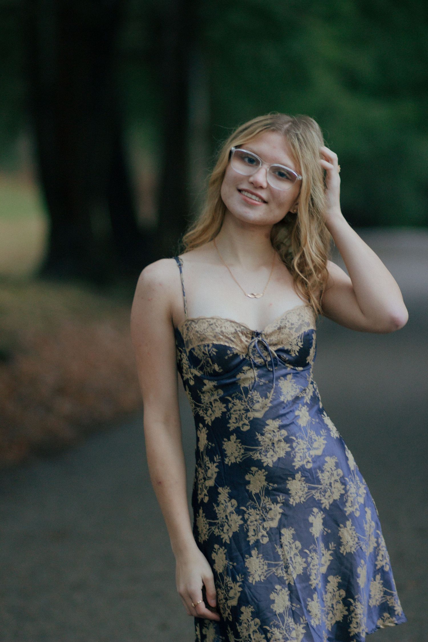 Young woman in floral blue dress and glasses smiling outdoors in a lush green park setting.