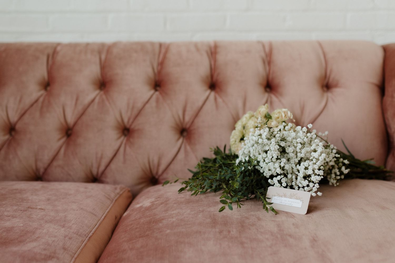 White floral bouquet with greenery resting on a pink tufted velvet sofa.