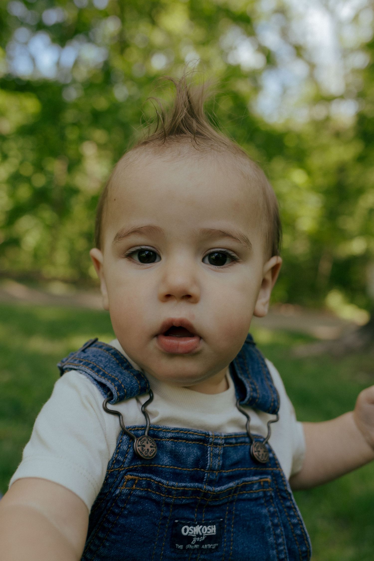Close-up portrait of a cute baby in denim OshKosh overalls with a tiny mohawk outdoors.