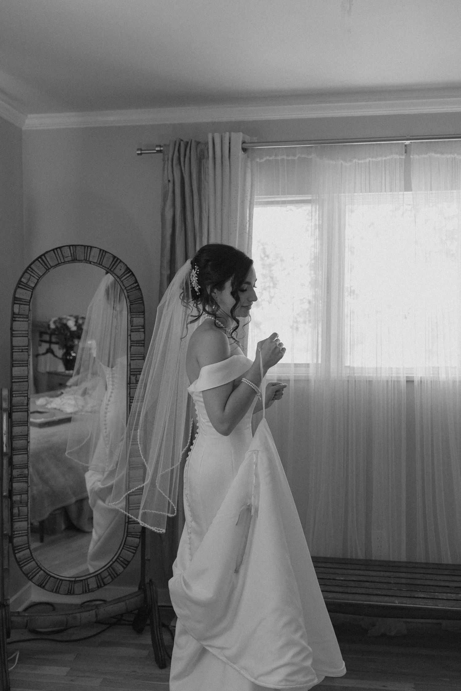 Black and white portrait of bride in wedding gown and veil standing by window with mirror reflection.