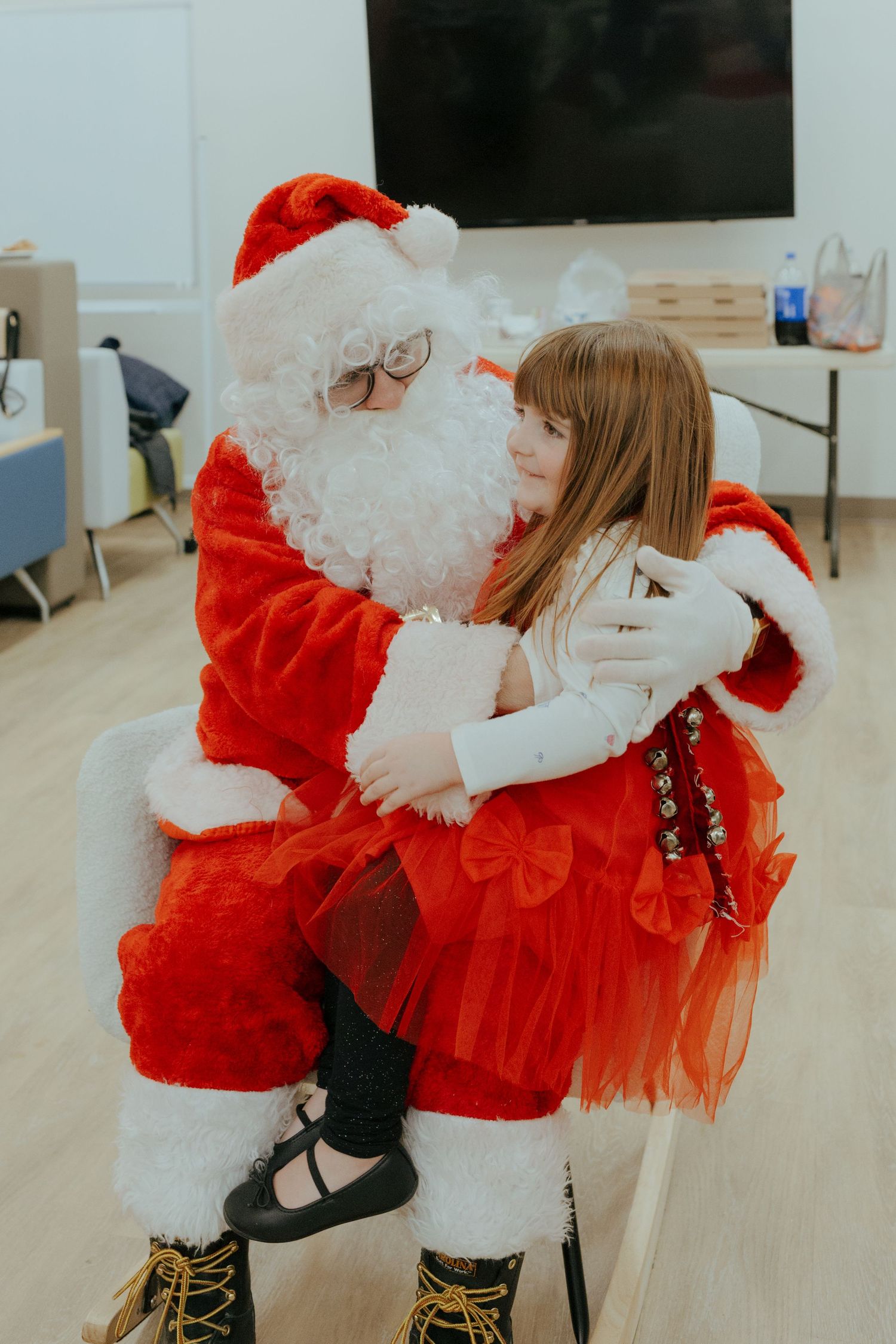 A young girl in a red tulle dress sits on Santa Claus's lap sharing a warm moment at a Christmas event.