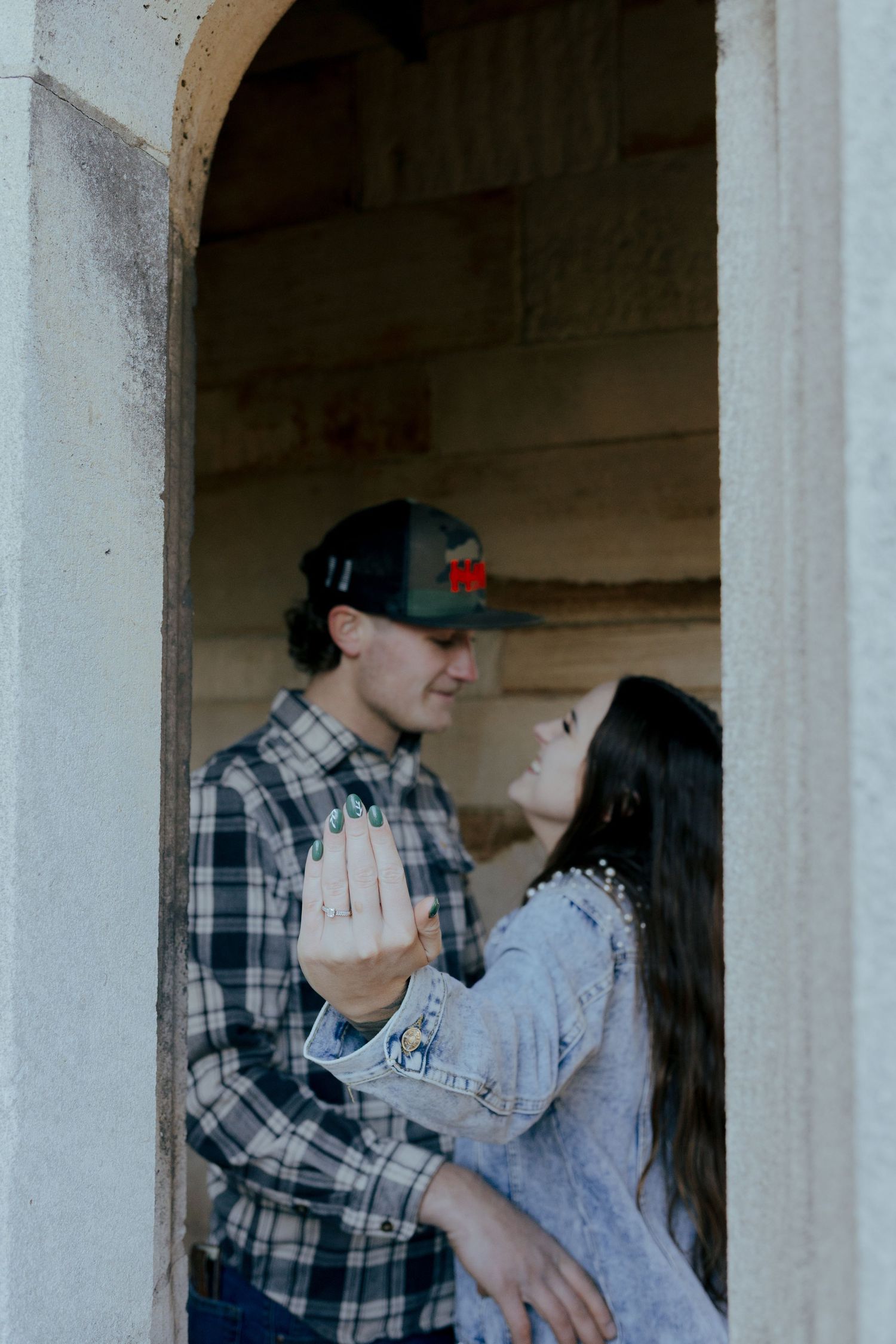 Engaged couple embraces in stone archway as woman shows off diamond engagement ring.