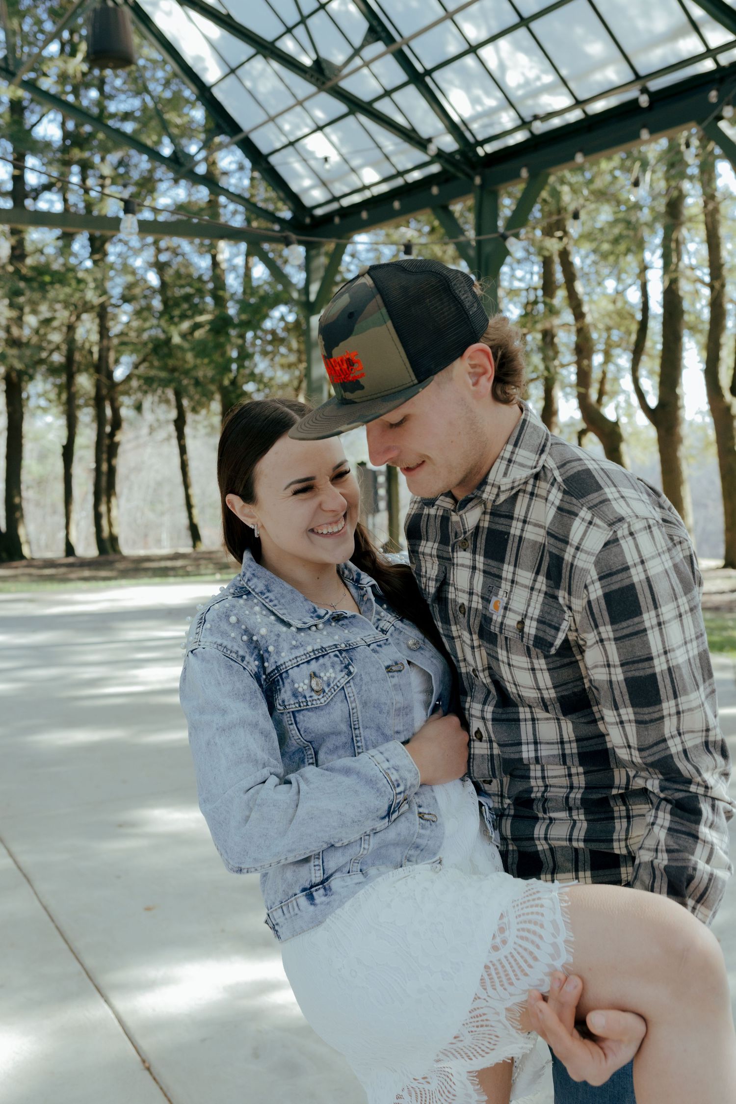 Smiling couple sits closely together under a glass pavilion surrounded by trees.