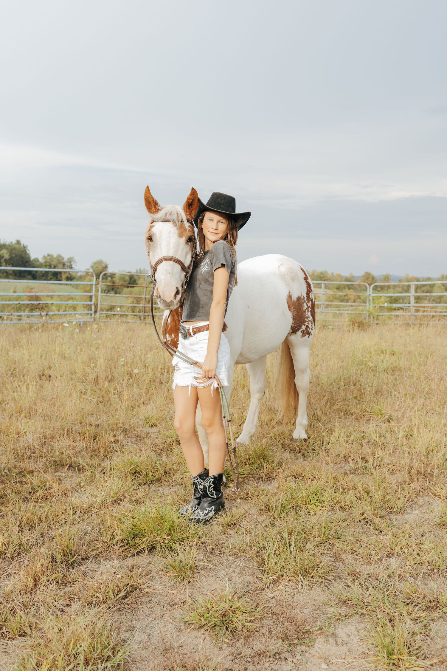 Young girl in black cowboy hat stands with a paint horse in a pasture.
