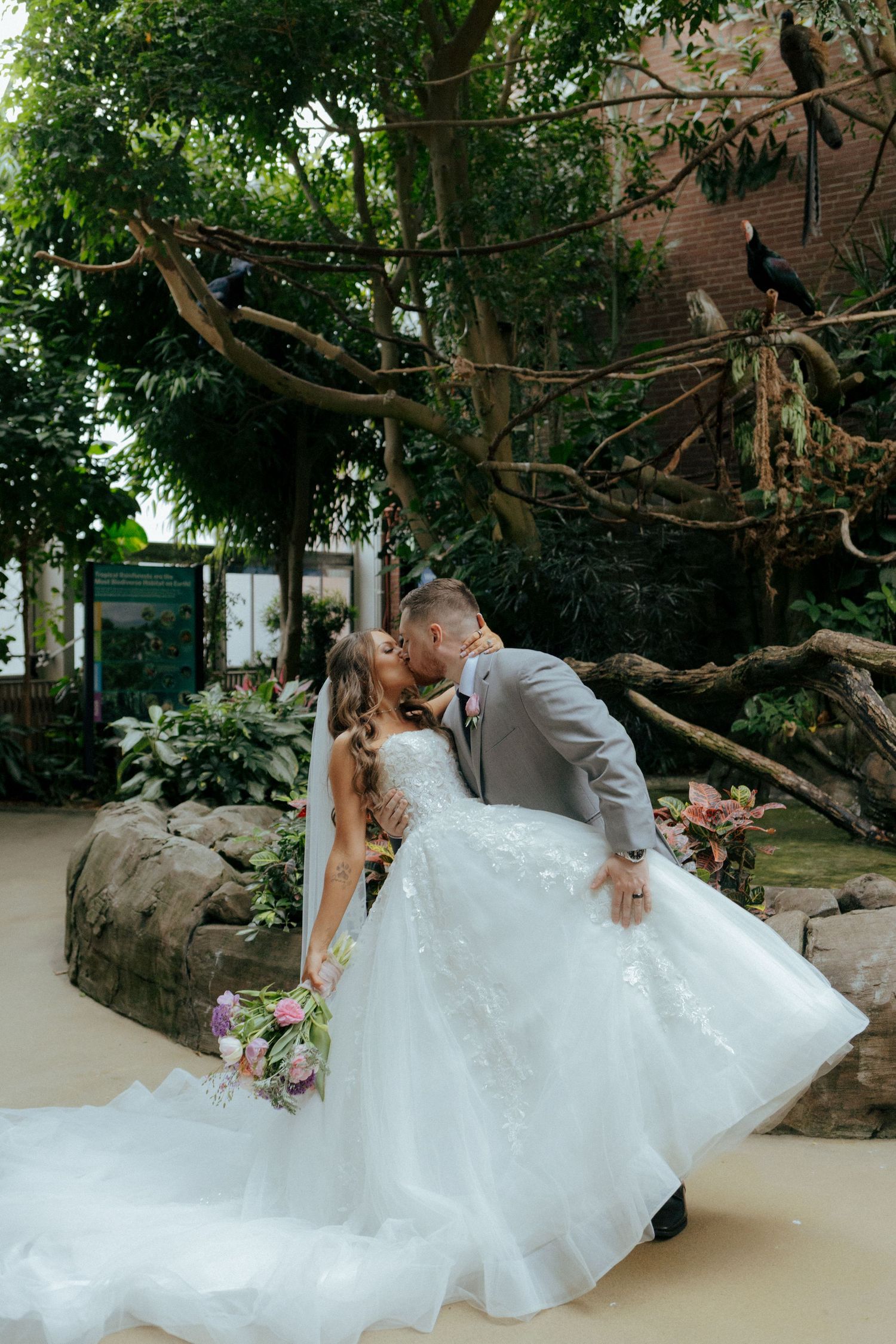 Bride and groom share a romantic kiss in a lush tropical garden setting, with birds perched in trees above them.