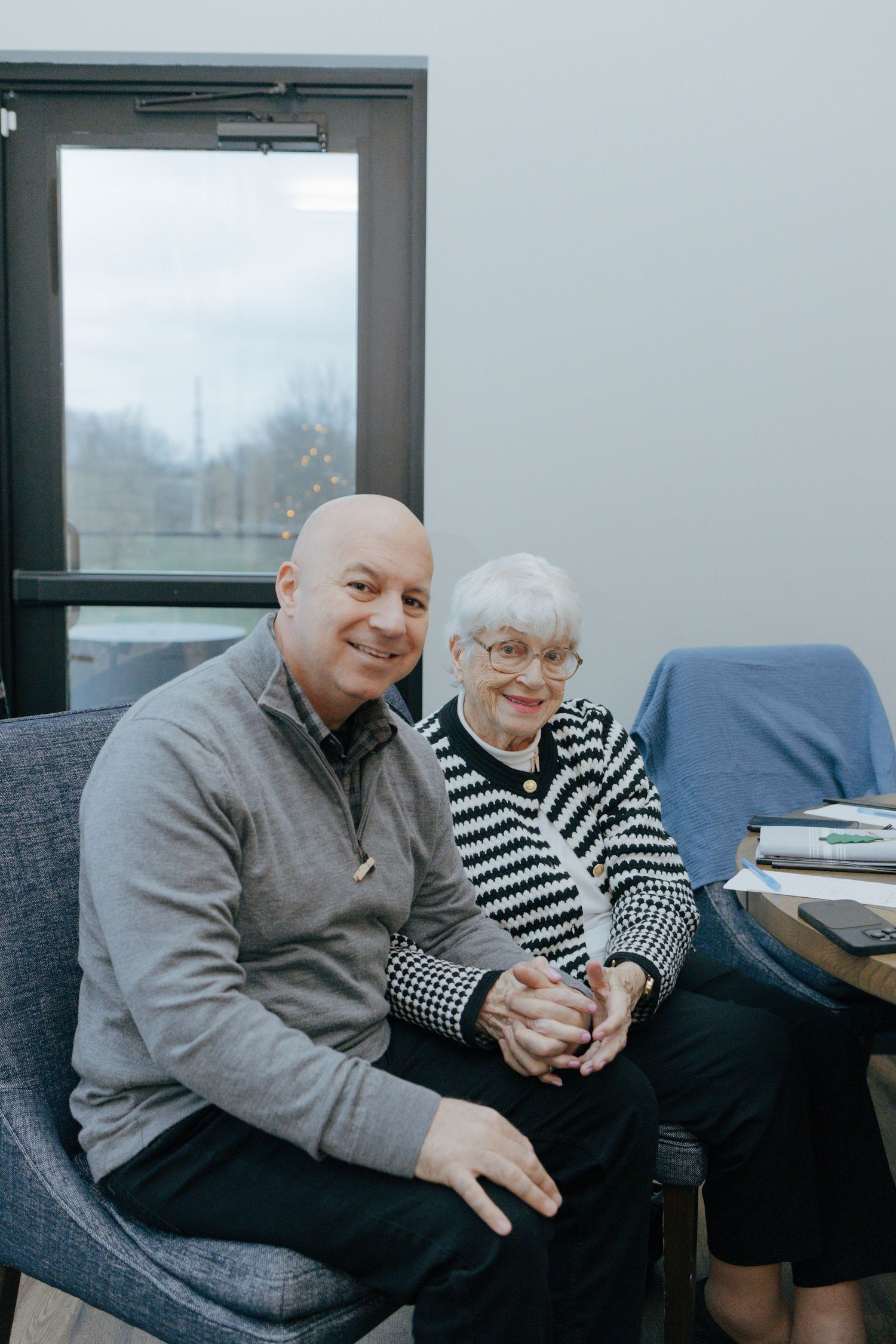 A bald man in a gray sweater sits smiling beside an elderly woman in a striped cardigan in a modern indoor setting.