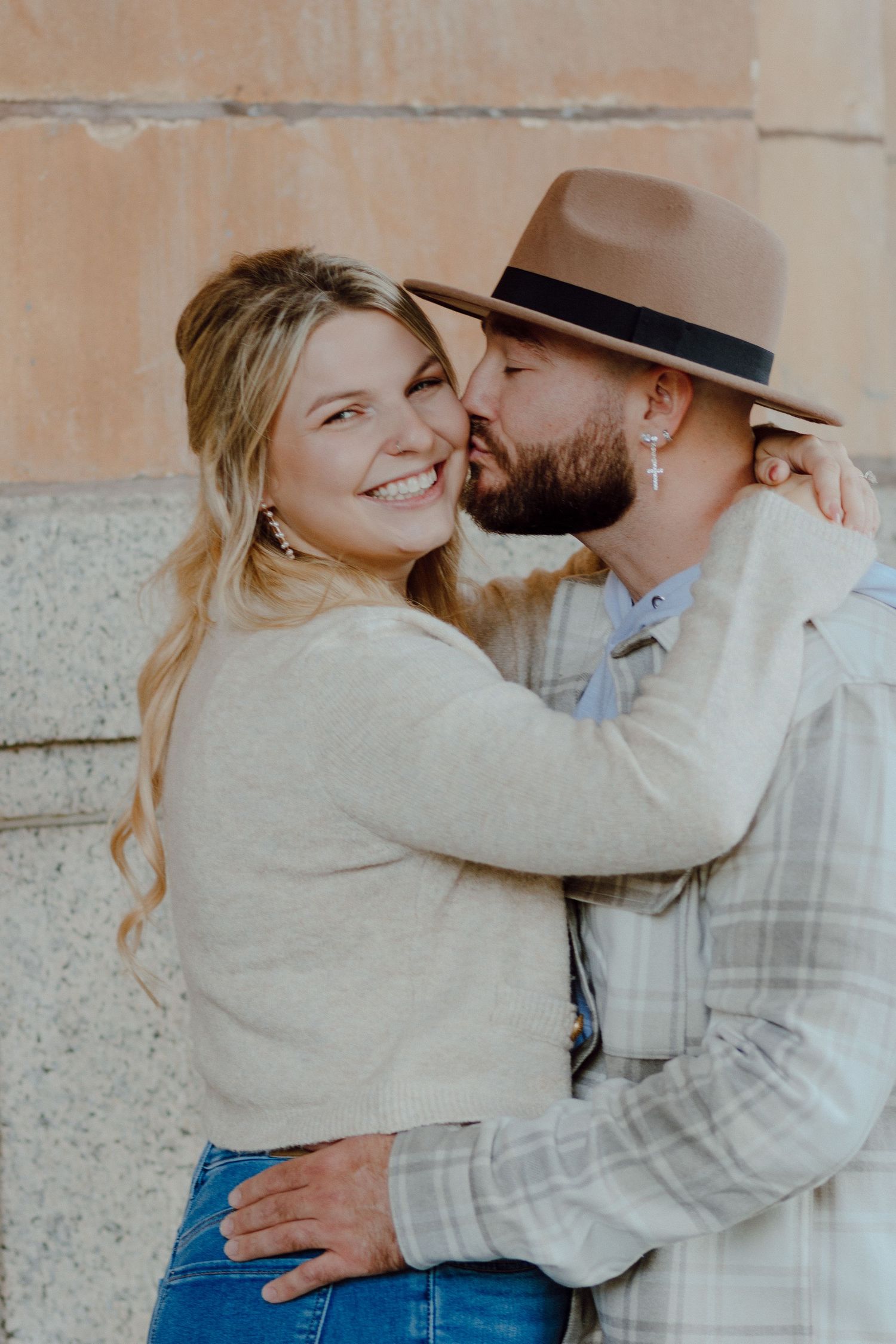 Bearded man in a brown fedora kissing the cheek of his smiling blonde fiancee against a stone wall backdrop.