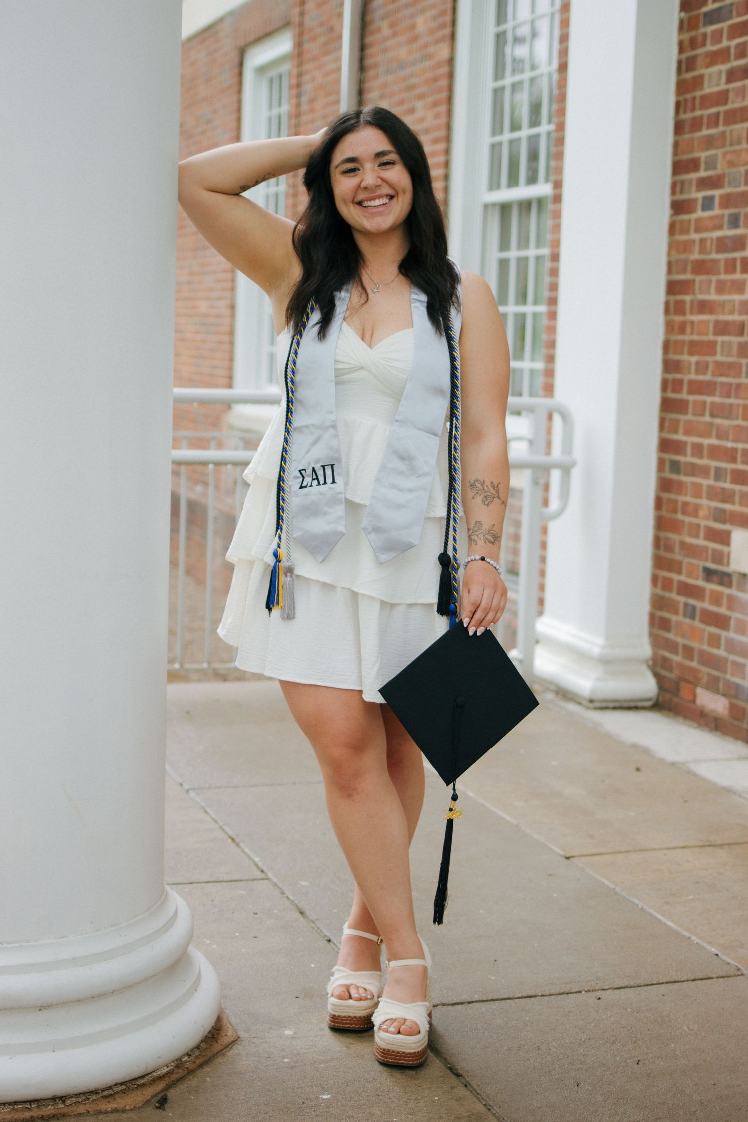 A smiling graduate in a white dress holds her cap while wearing a sorority stole outside a brick building at Slippery Rock