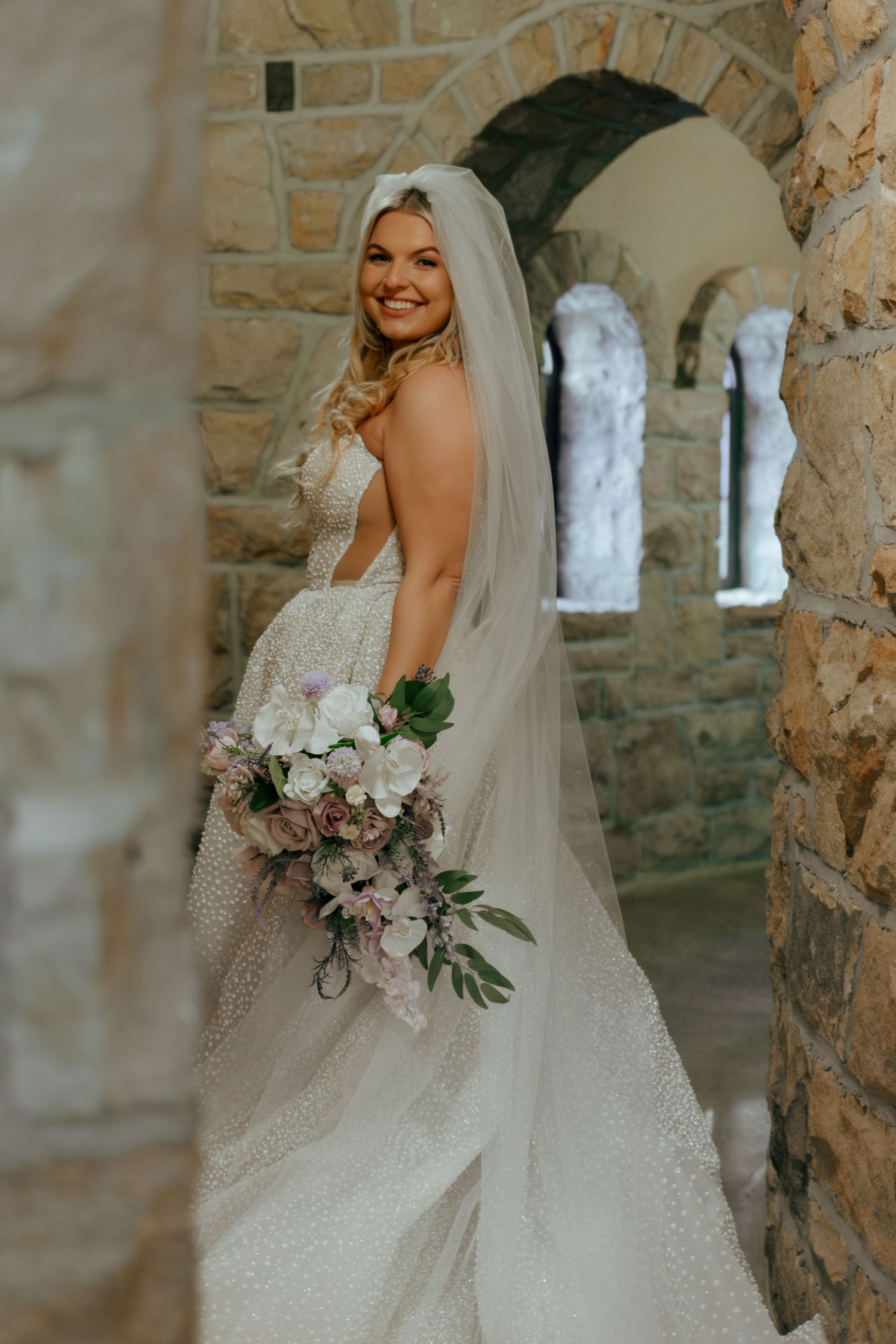 Beautiful bride in a pearl-embellished gown holding a lavender bouquet while posing in a stone archway.