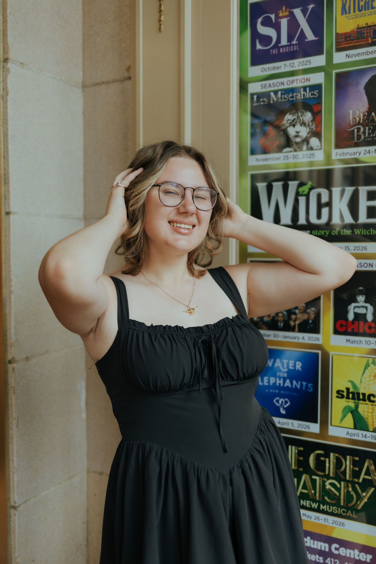 Smiling young woman in black dress poses before Broadway show posters including Wicked and Six.