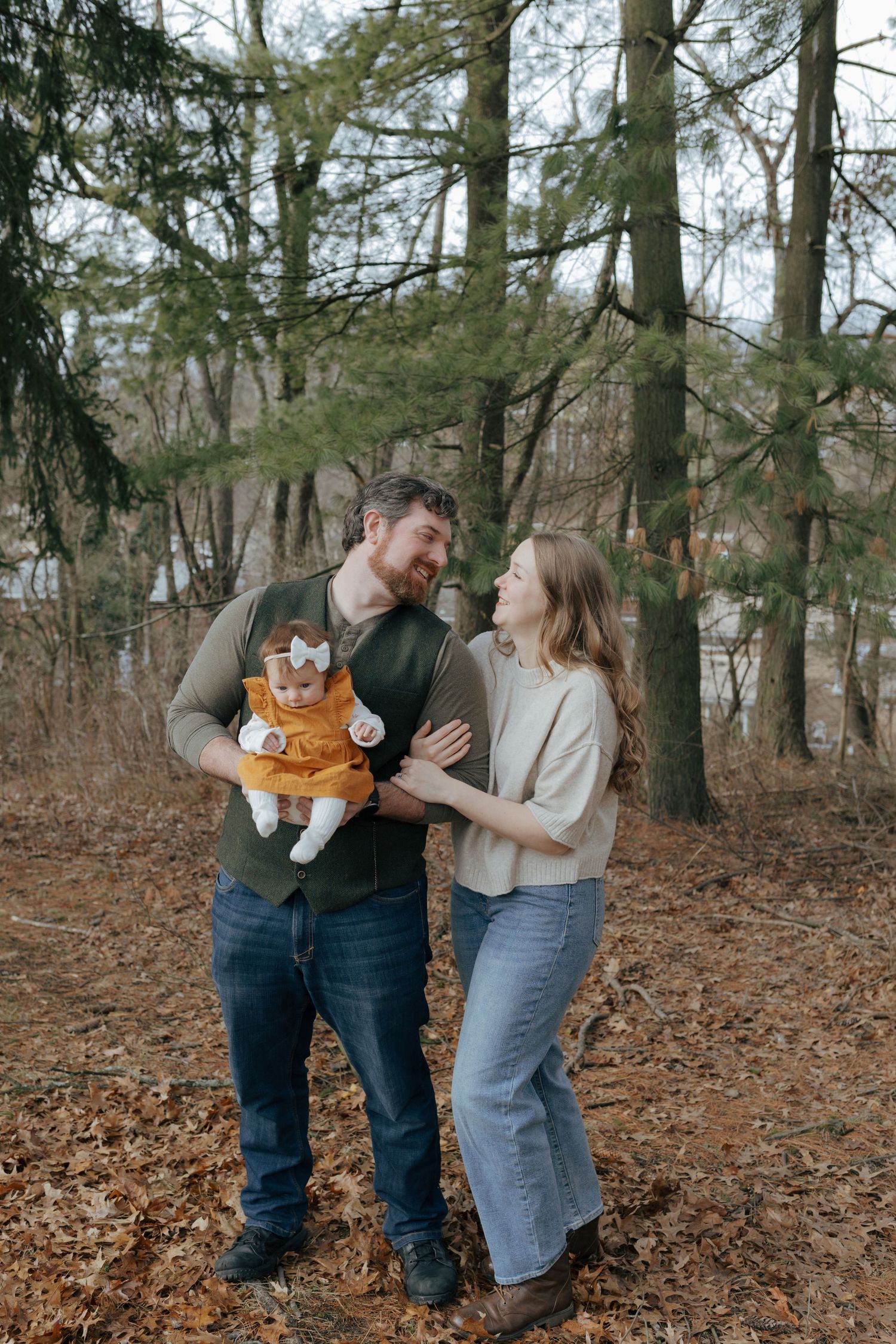 A happy young family with a baby in an orange outfit poses together in an autumn forest setting.