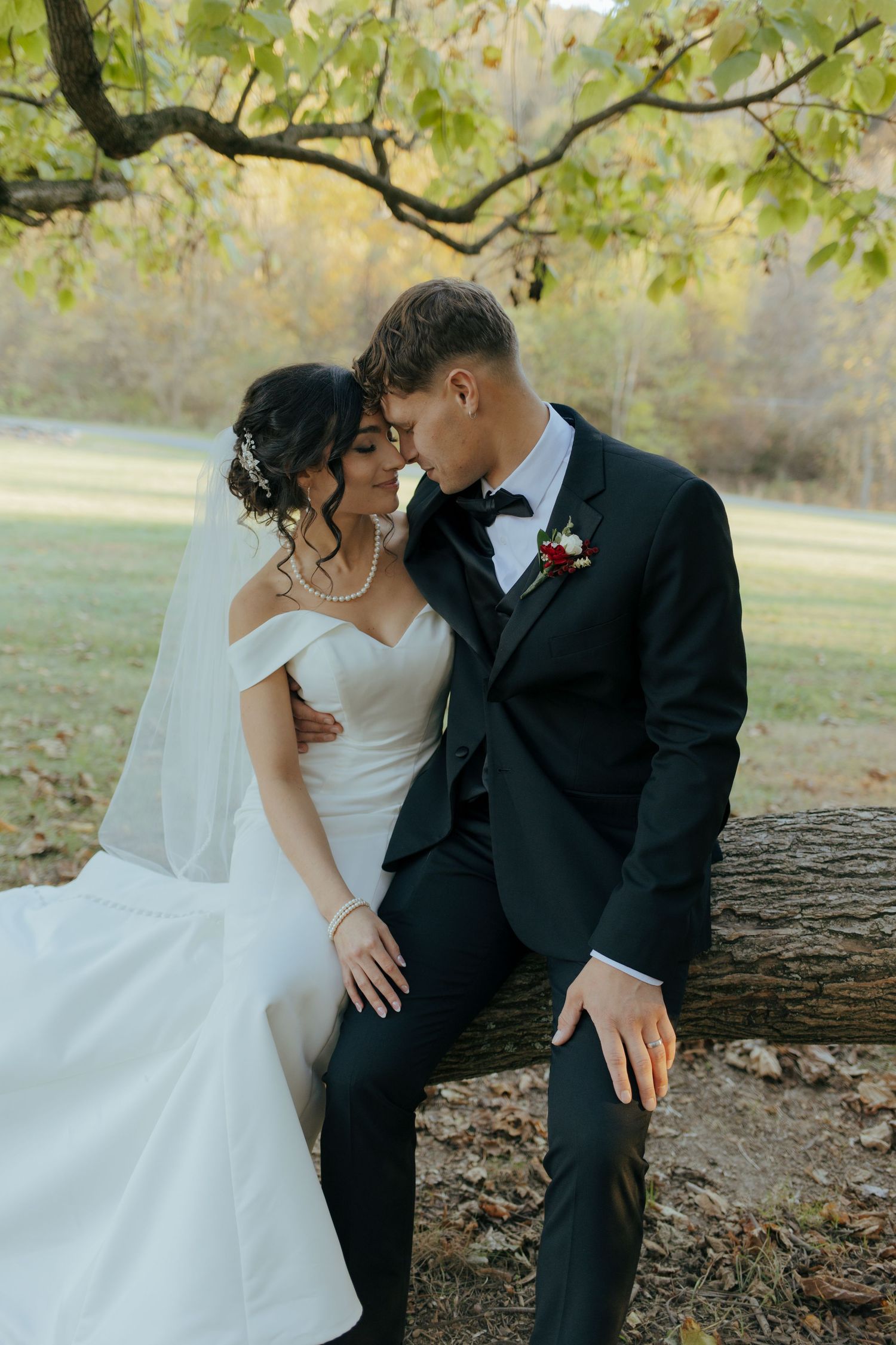 Bride and groom share romantic moment seated on log under autumn trees in park.