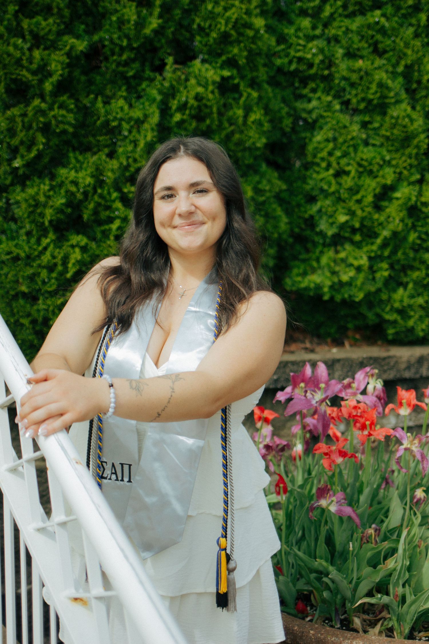 Smiling graduate in white dress leans on white railing beside vibrant tulips and green hedges.