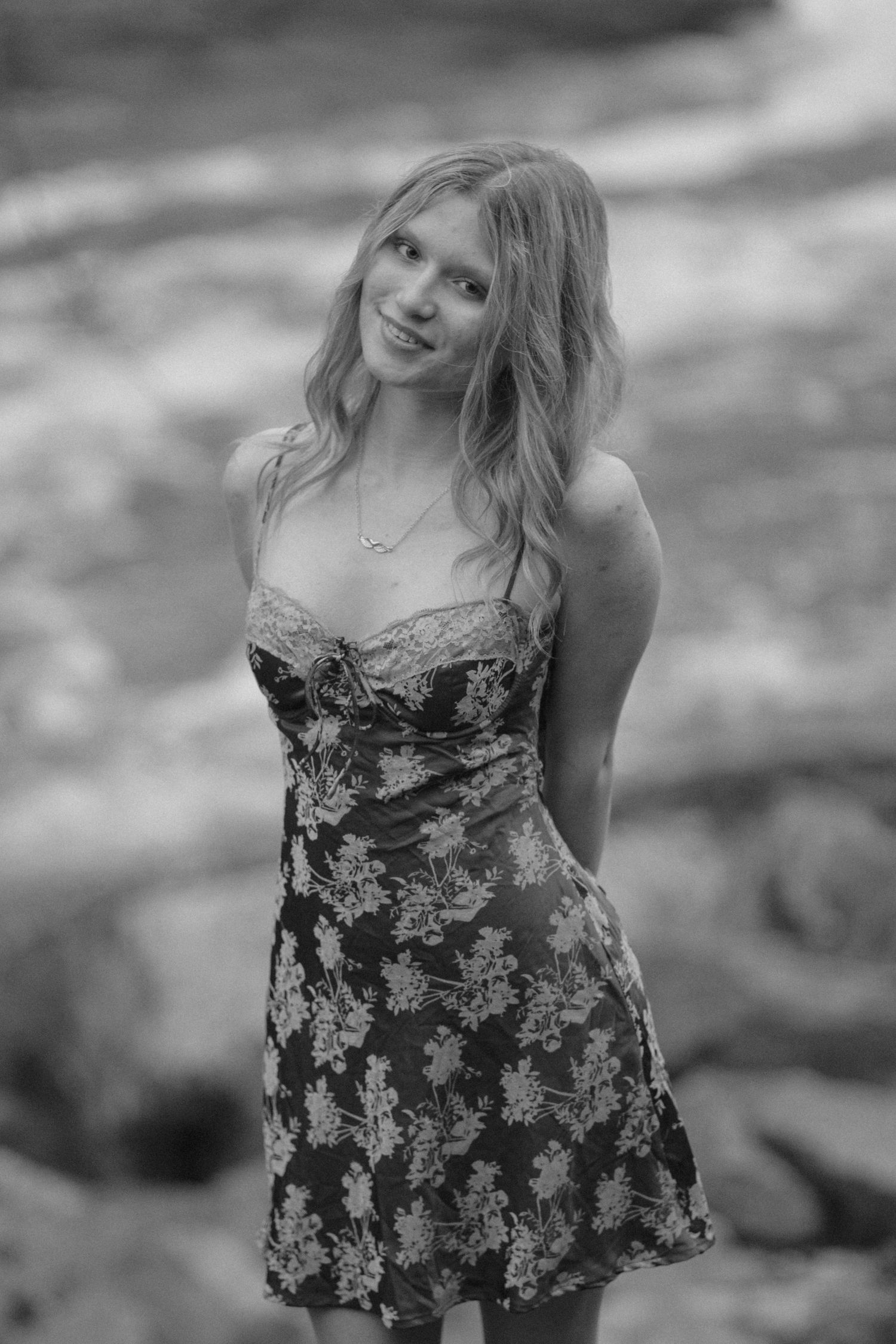 Black and white portrait of a smiling blonde teen in a floral dress near a rocky stream.