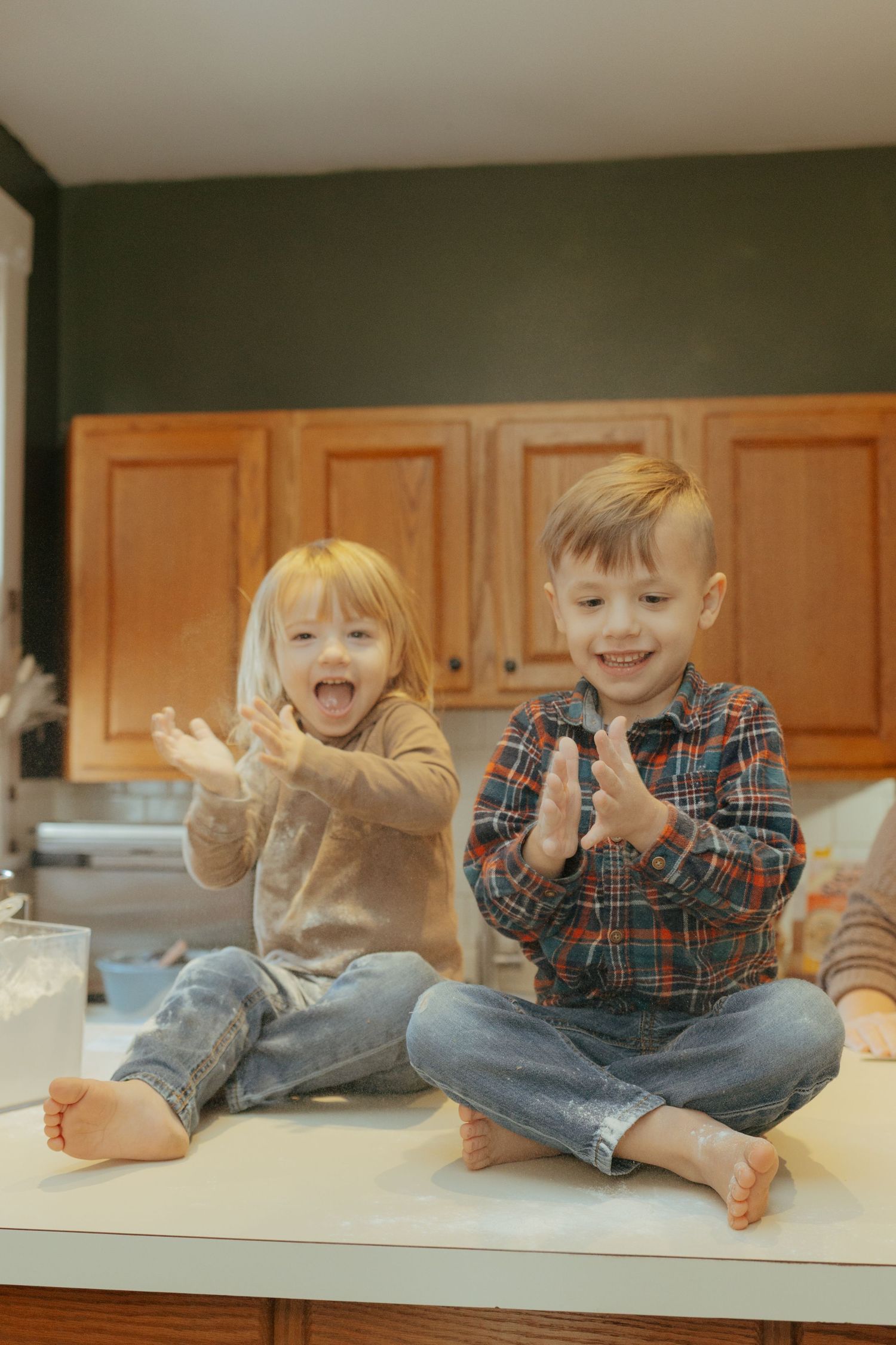Two young siblings clapping flour-covered hands together laughing while sitting on kitchen counter.