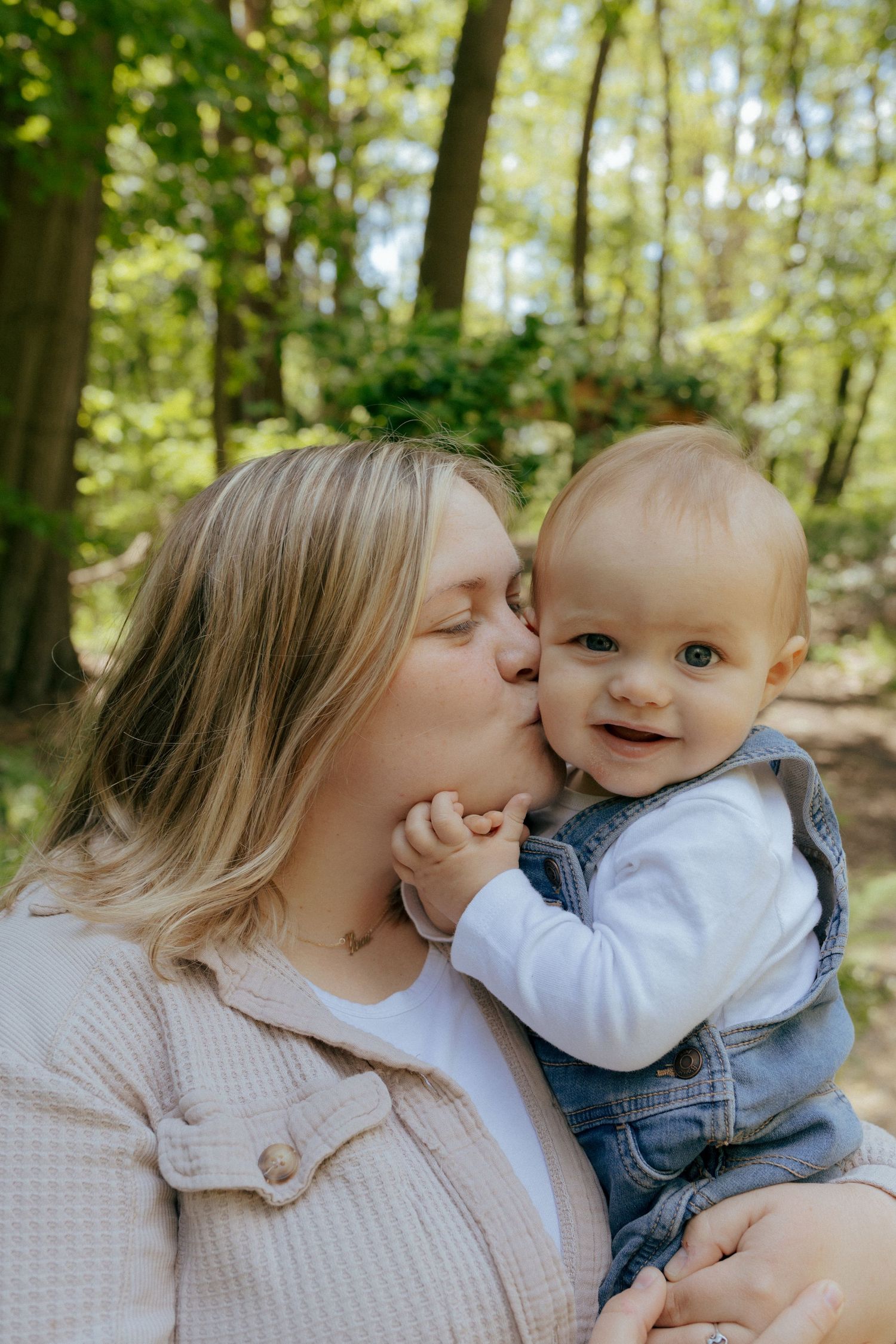 Blonde mother kissing her smiling baby in denim overalls in a sunlit forest setting.