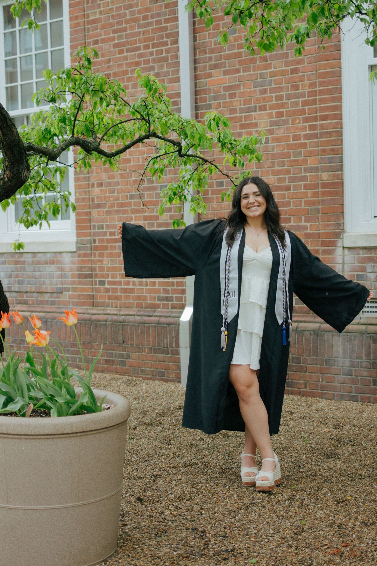 Joyful college graduate in black gown posing with open arms near brick building and tulips.