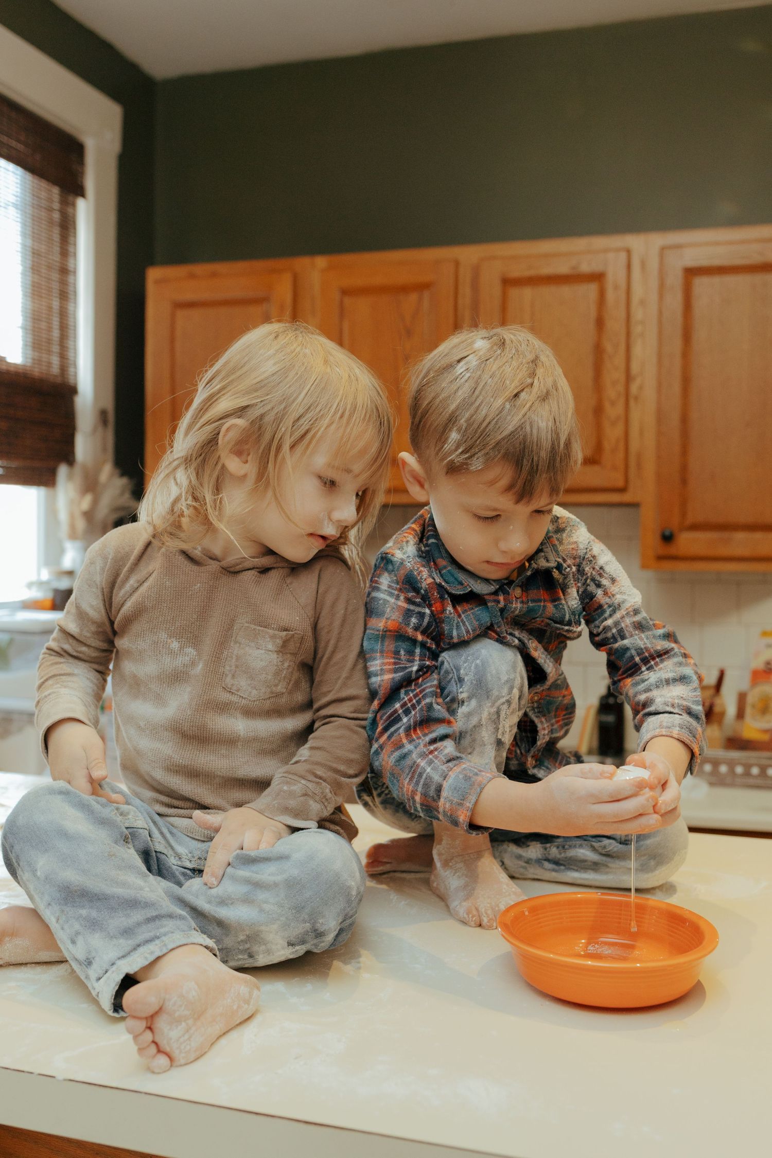 Two young siblings sitting on flour-dusted kitchen counter cracking eggs into an orange bowl.