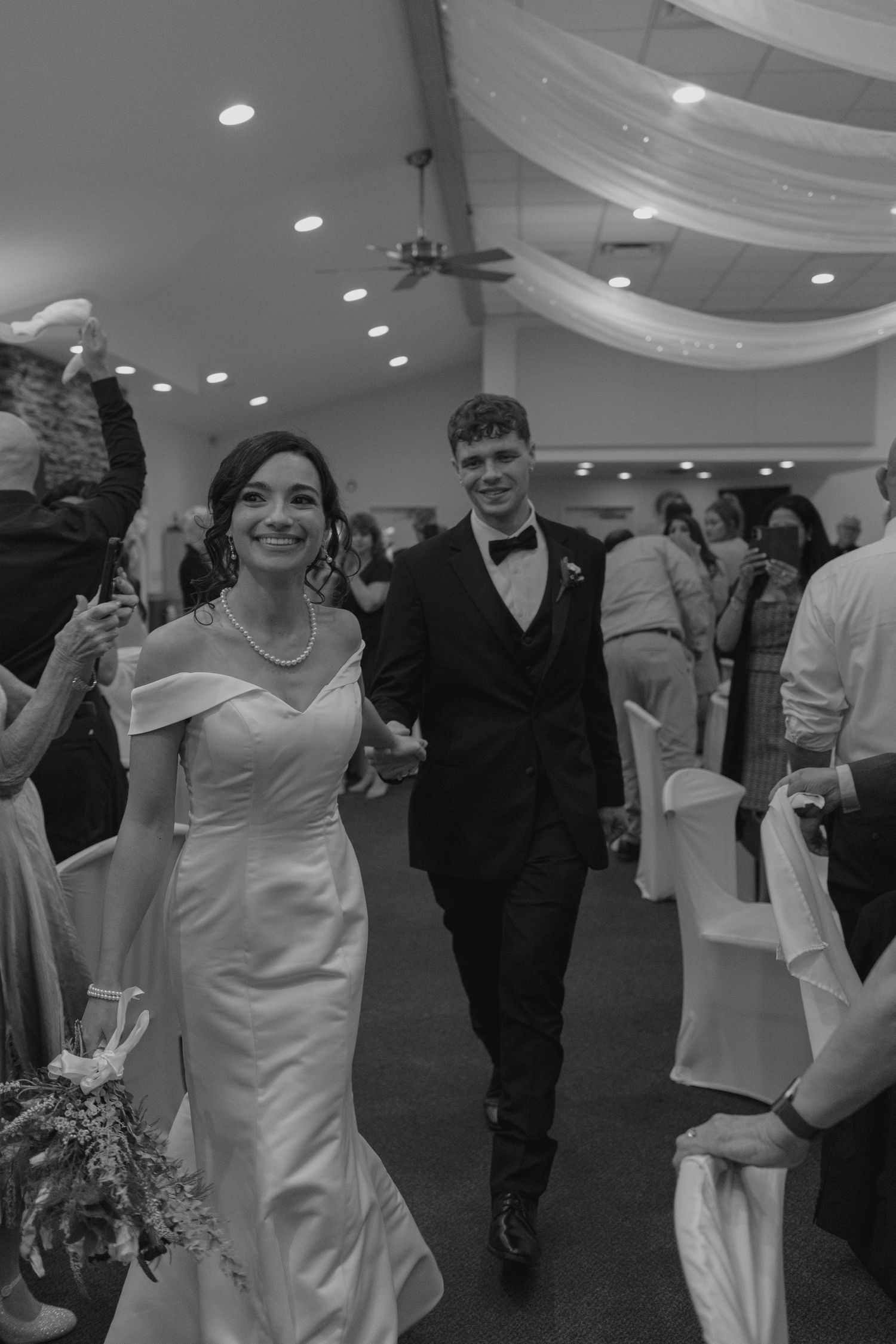 Black and white photo of smiling newlyweds walking through wedding reception hall guests.