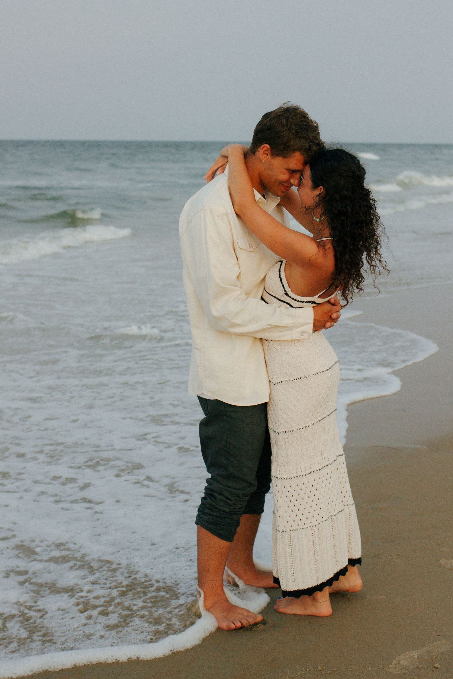 Couple embracing romantically on a beach shoreline with ocean waves crashing behind them.