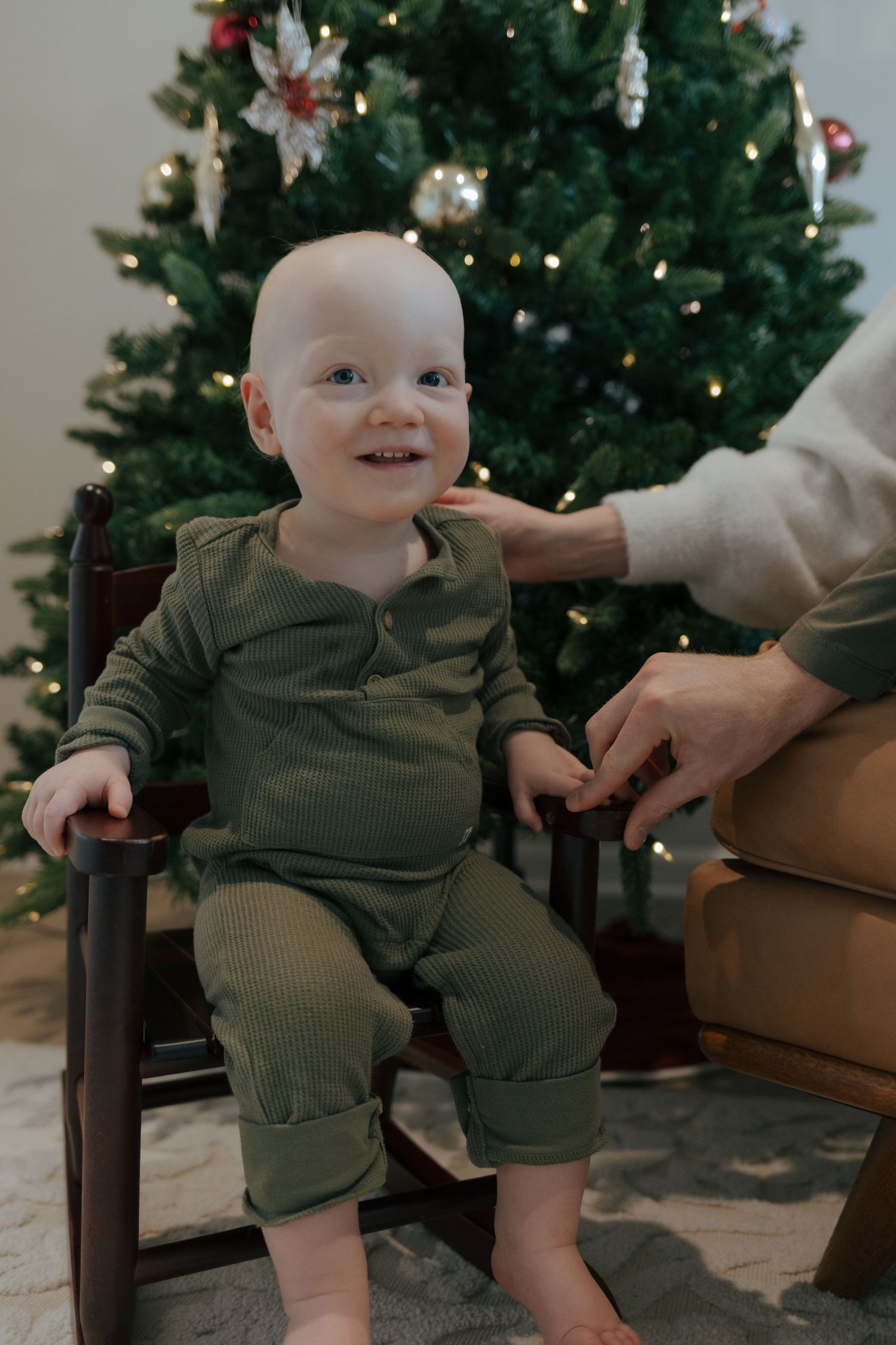 An adorable smiling baby in a green outfit sits in a chair near a decorated Christmas tree.