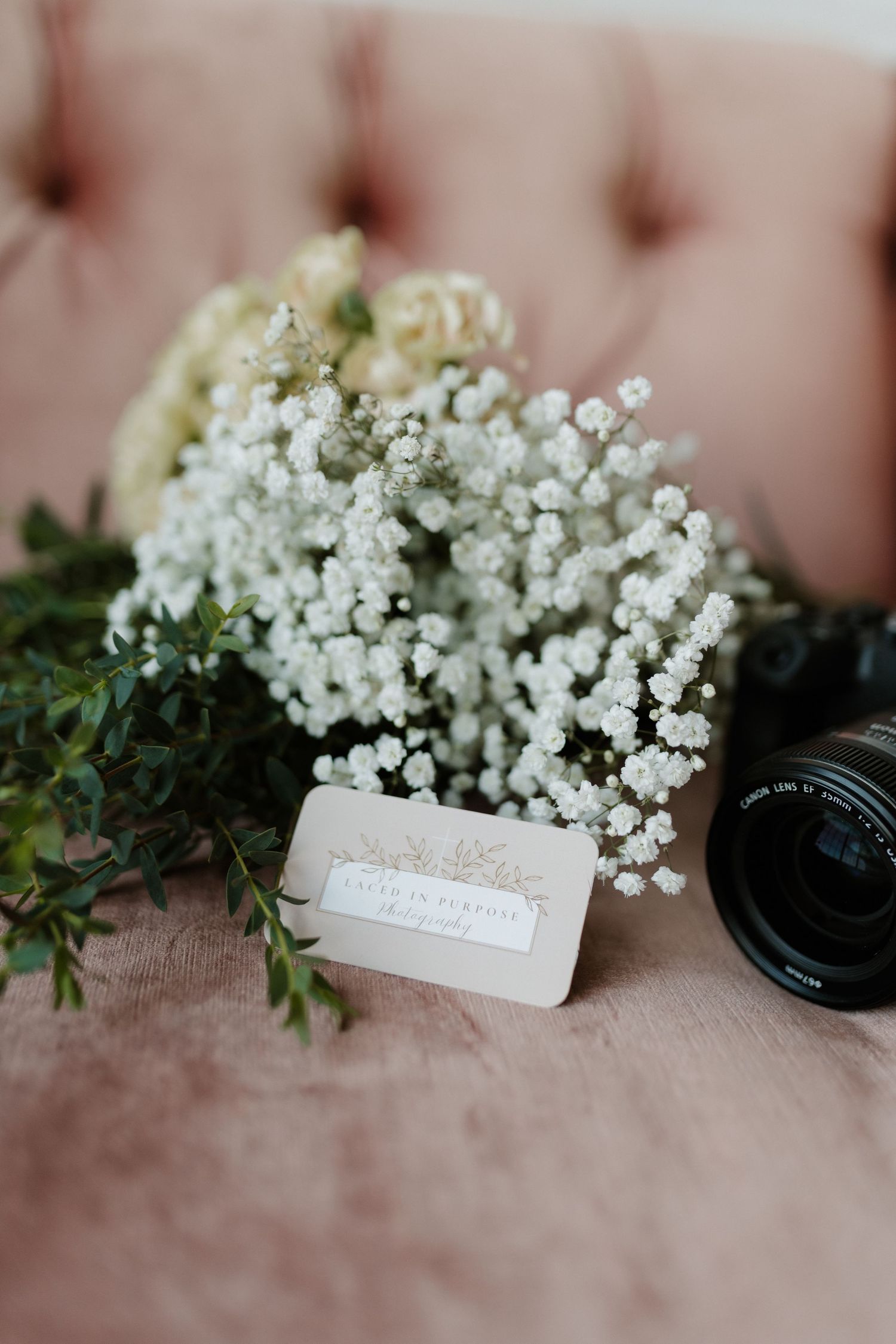 White floral bouquet with business card and camera lens on pink velvet sofa.