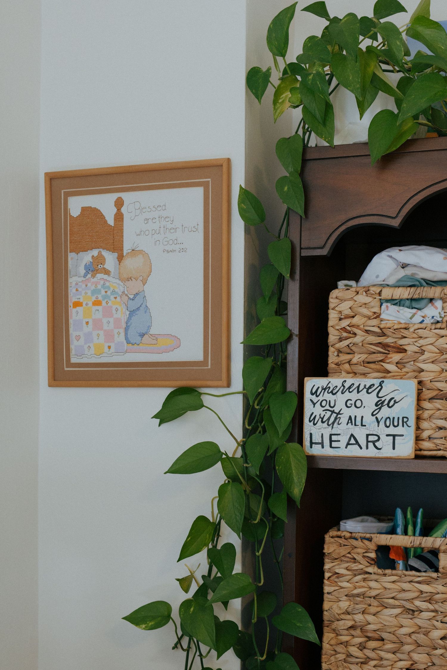 Nursery corner with framed prayer art, trailing pothos plant, and wicker baskets.
