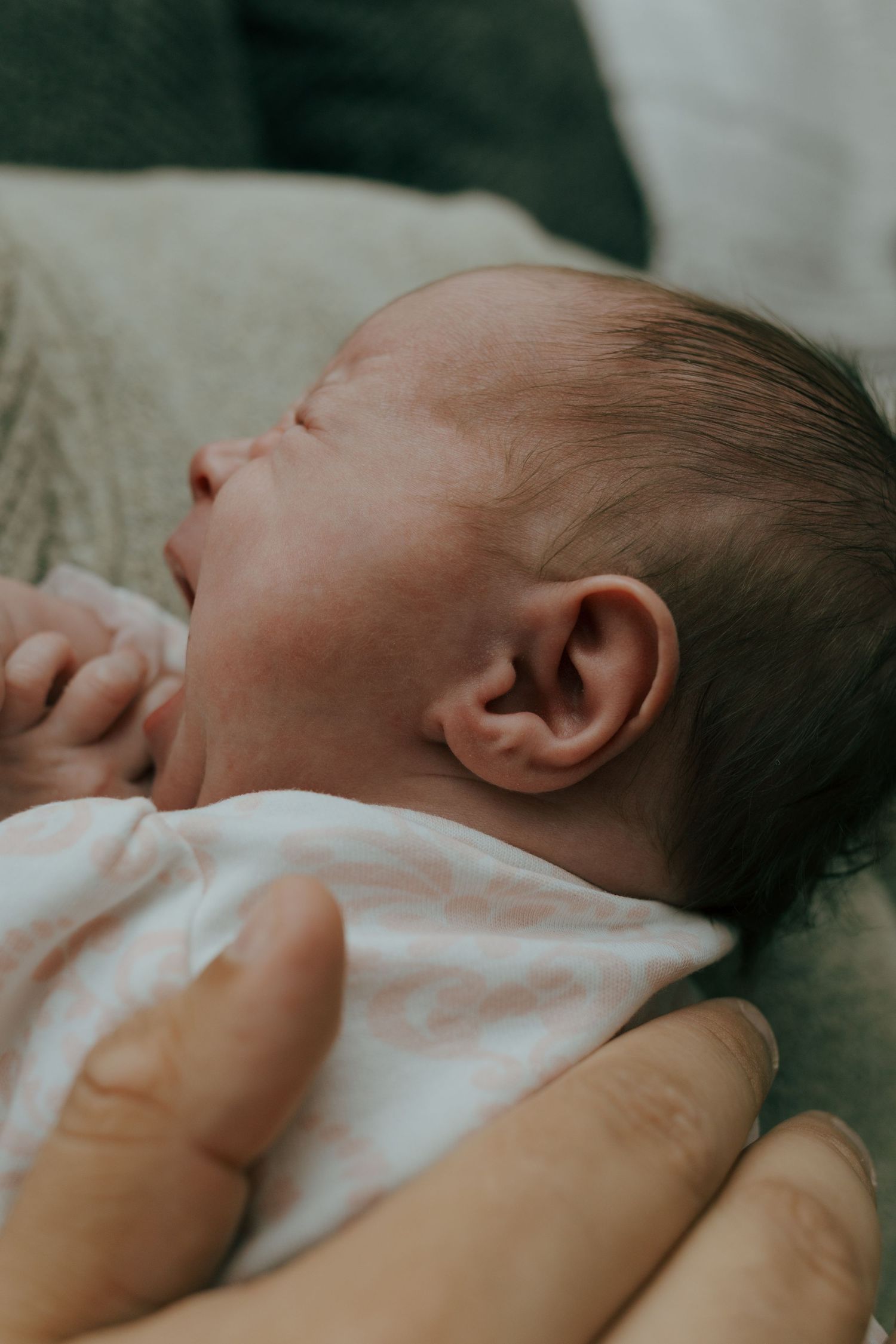 Peaceful newborn baby sleeping in parent's arms, wearing white onesie with tiny hands curled near face.