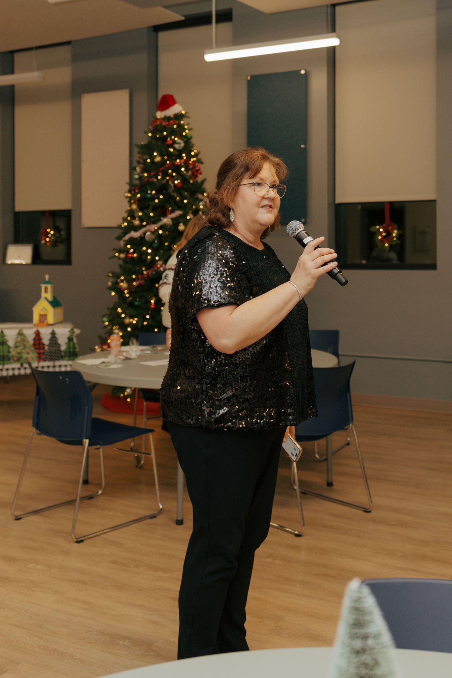 A woman in a sequined black top speaks into a microphone in front of a decorated Christmas tree at a holiday event.