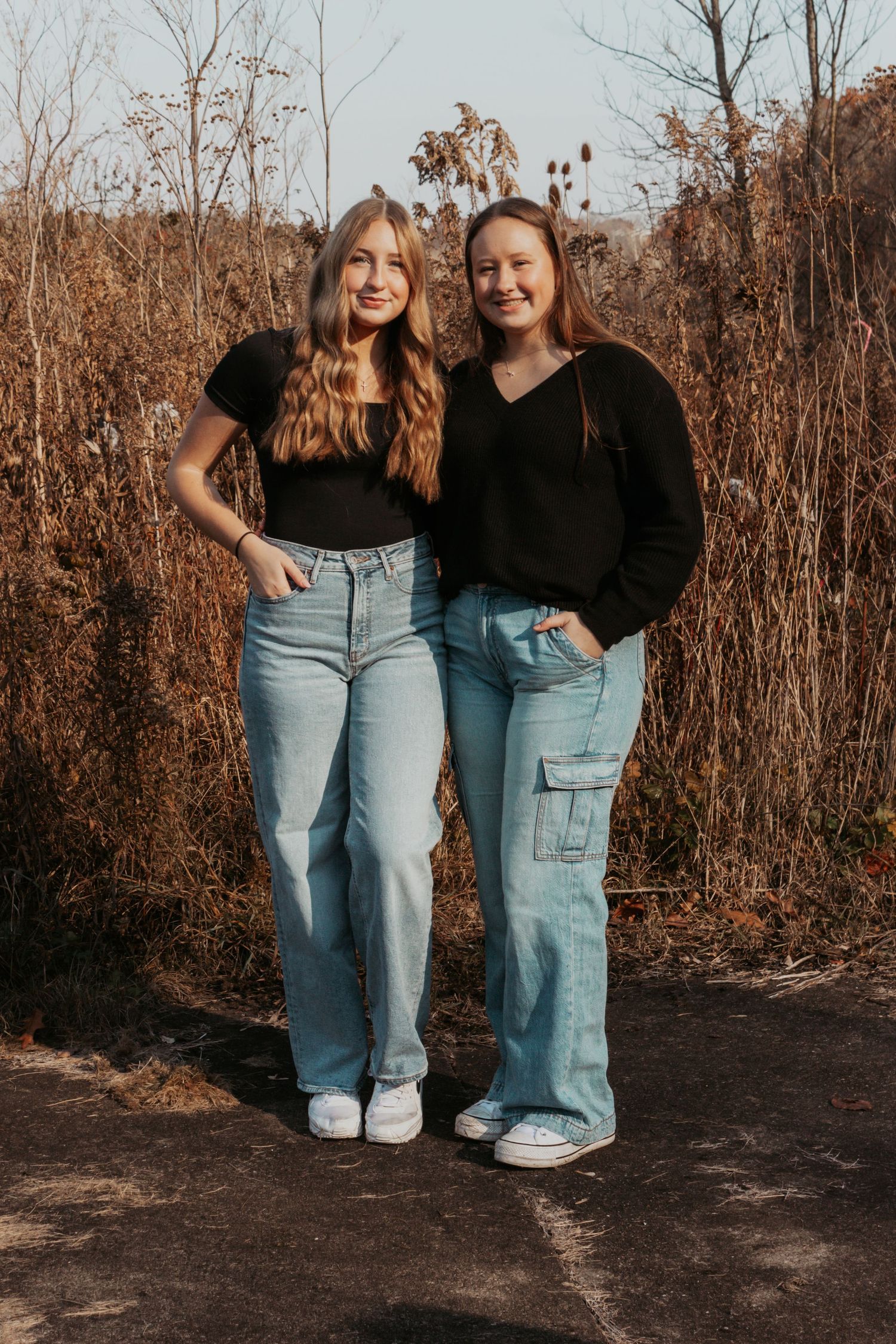 Two young women in black tops and light jeans smiling outdoors in fall setting.