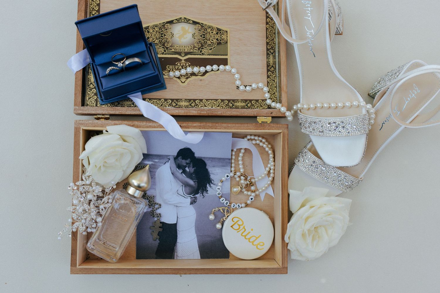 Elegant wedding flat lay with rings, pearl jewelry, bridal shoes, white roses, and a romantic couple photo in a wooden box.