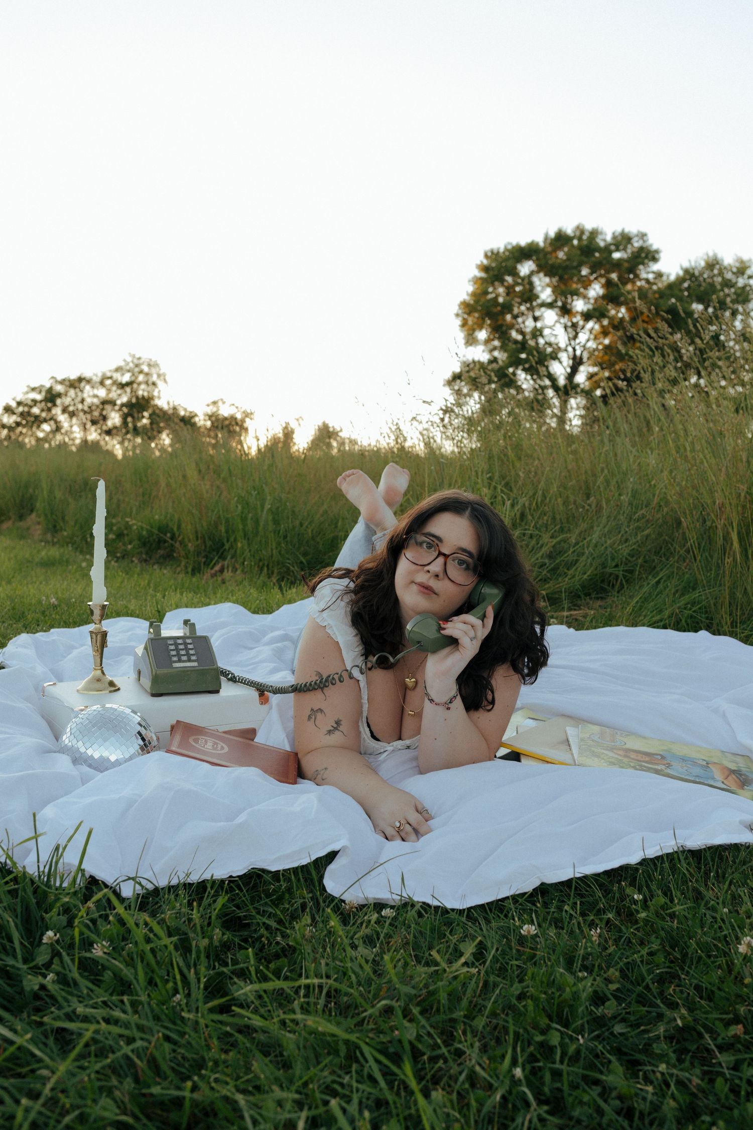 Young woman with glasses lying on white blanket in grass, talking on vintage phone with candle and crystal ball nearby.
