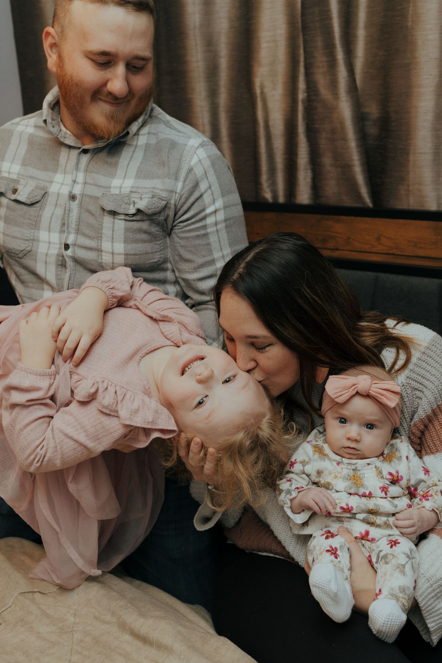 Loving mother kisses toddler daughter while father smiles and baby sits nearby on a couch.
